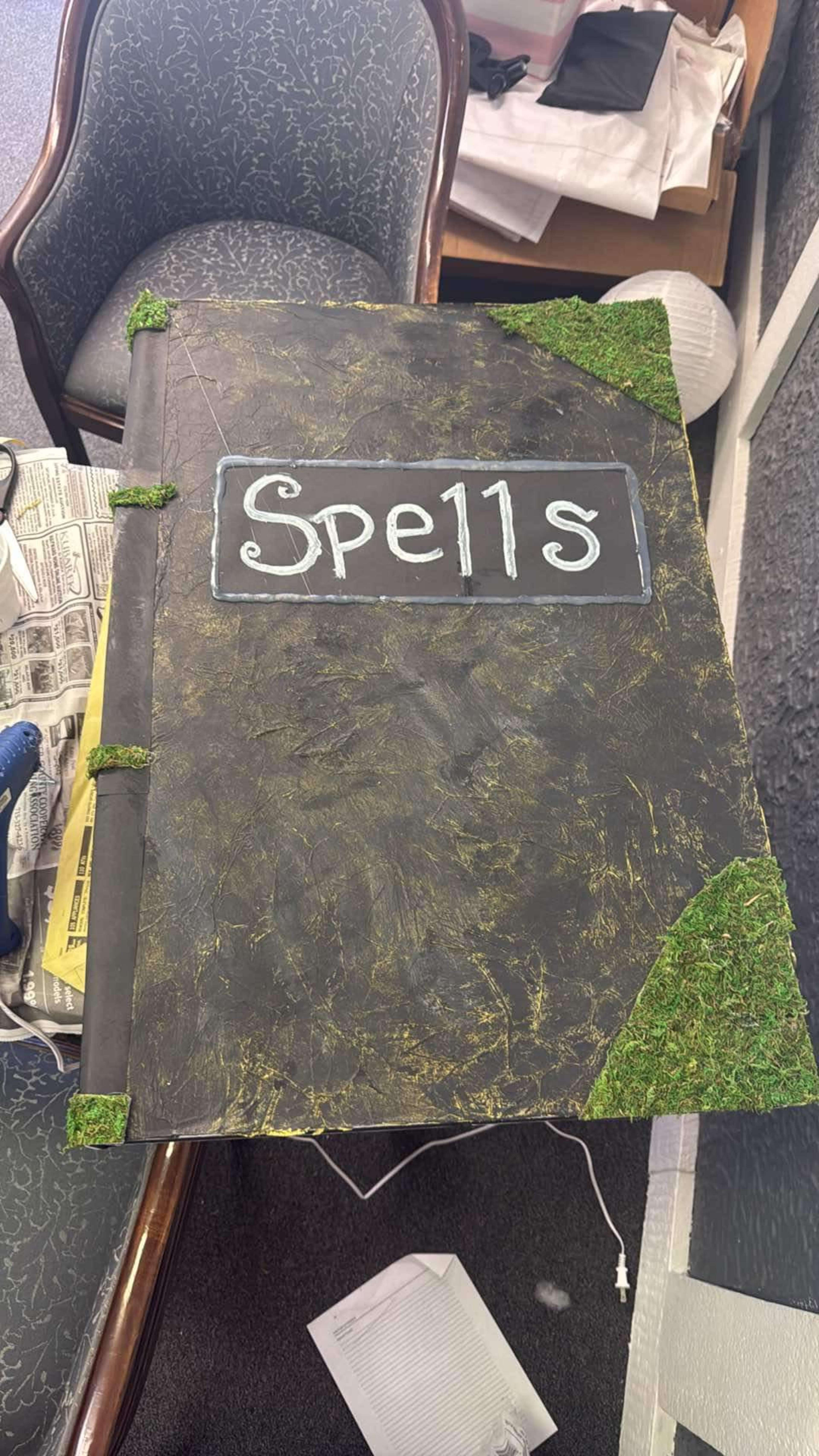 The image shows a large, ornate spellbook with a textured cover, decorated with moss-like greenery and the title "Spells" written in white on a black background.