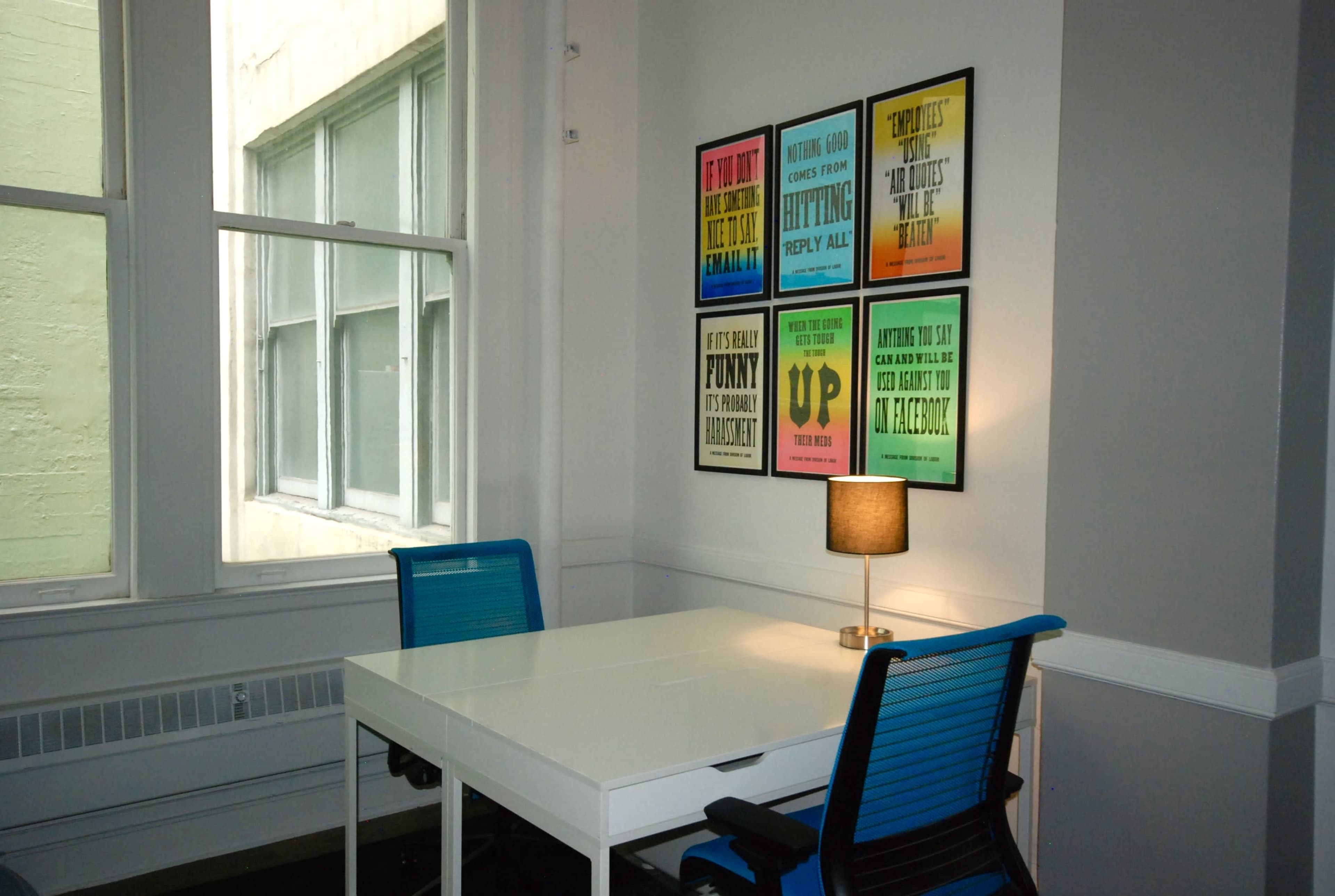 The image shows a small office space featuring a white desk with two blue chairs, illuminated by a lamp, and adorned with colorful framed posters on the wall.