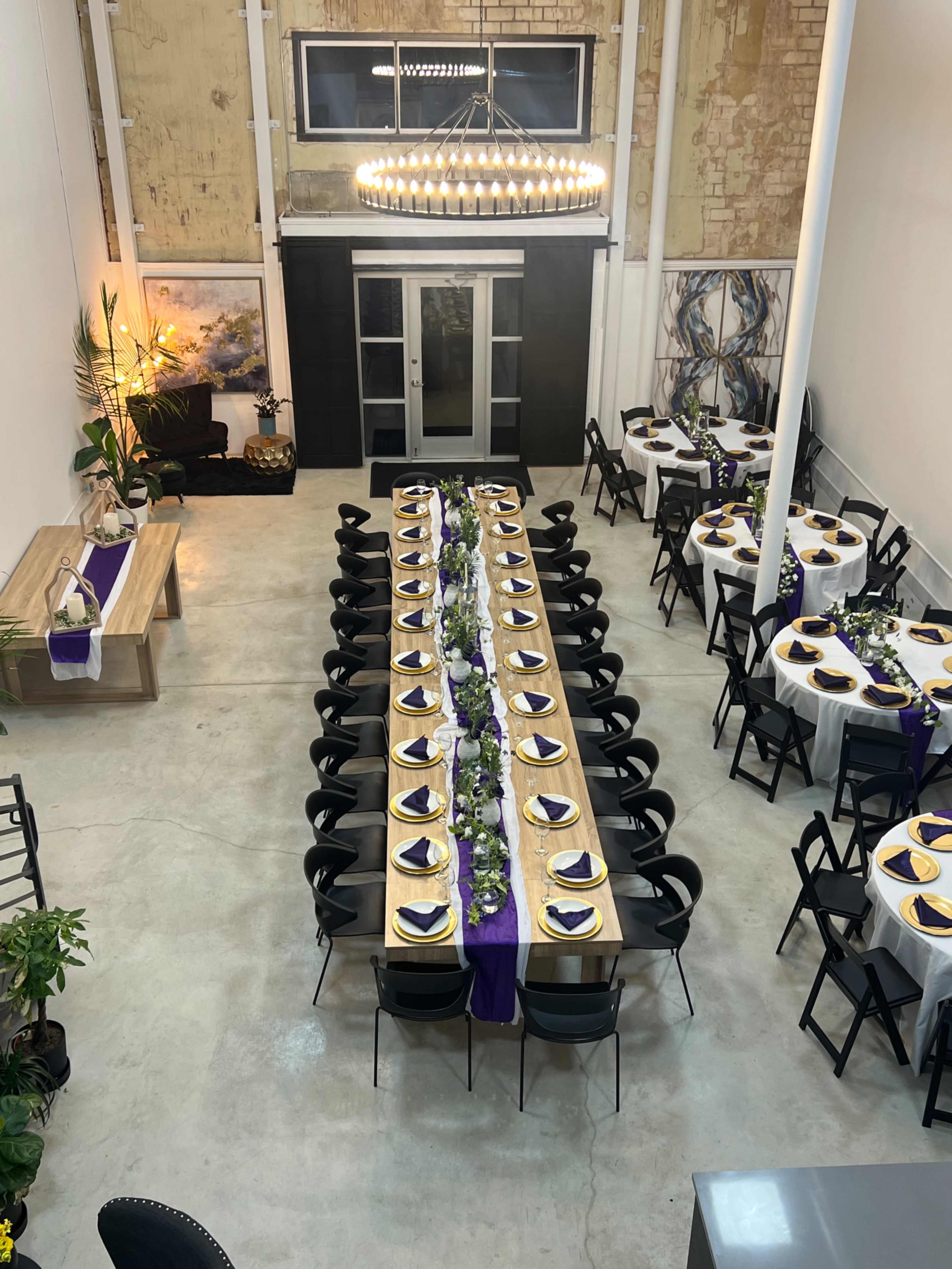 A large dining area features two long tables set with white tablecloths and purple accents, arranged for a formal event in a spacious room with exposed brick walls.