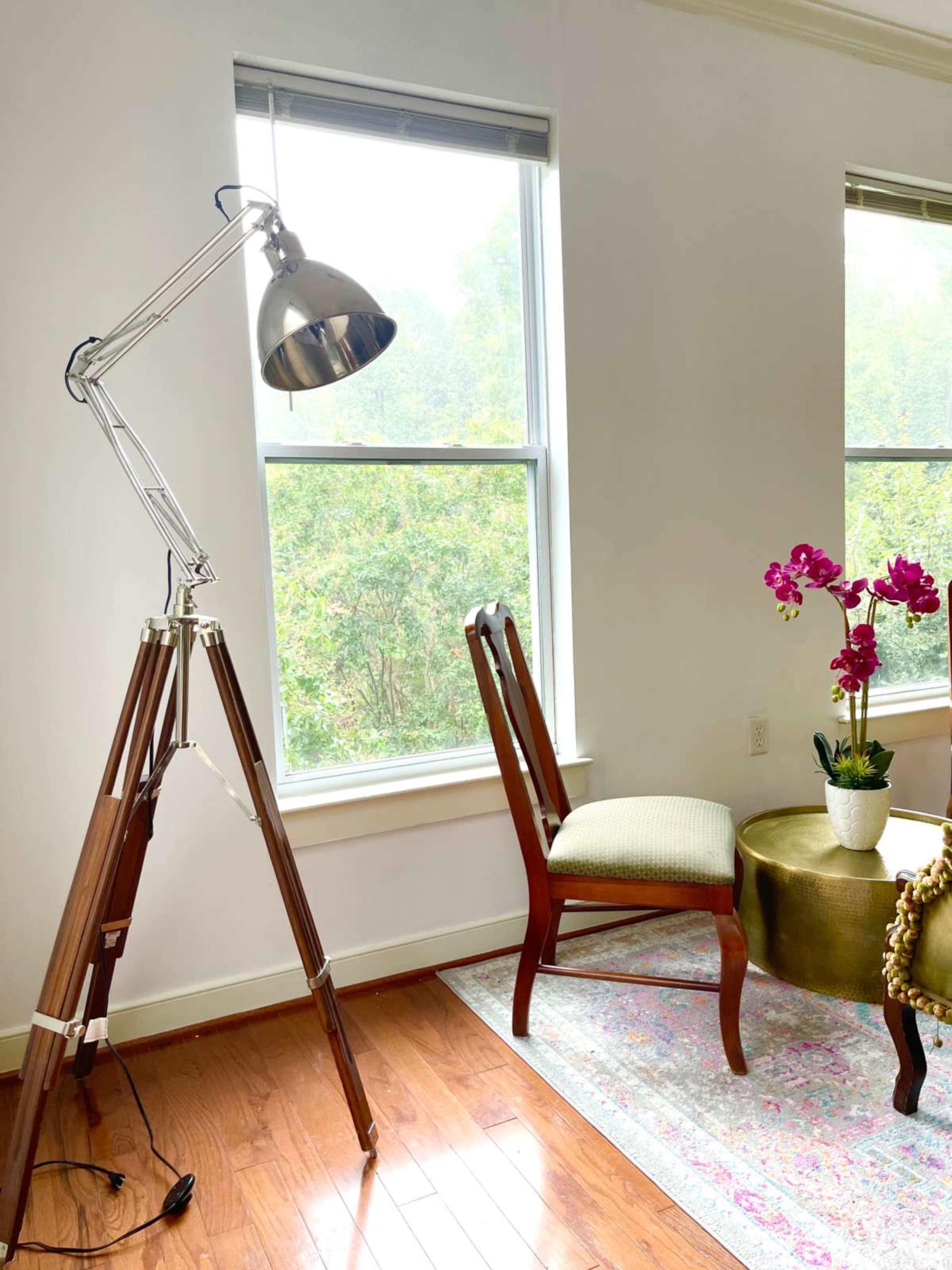 A polished metal floor lamp stands beside a wooden chair and a round table adorned with a potted orchid, illuminated by natural light from the windows.