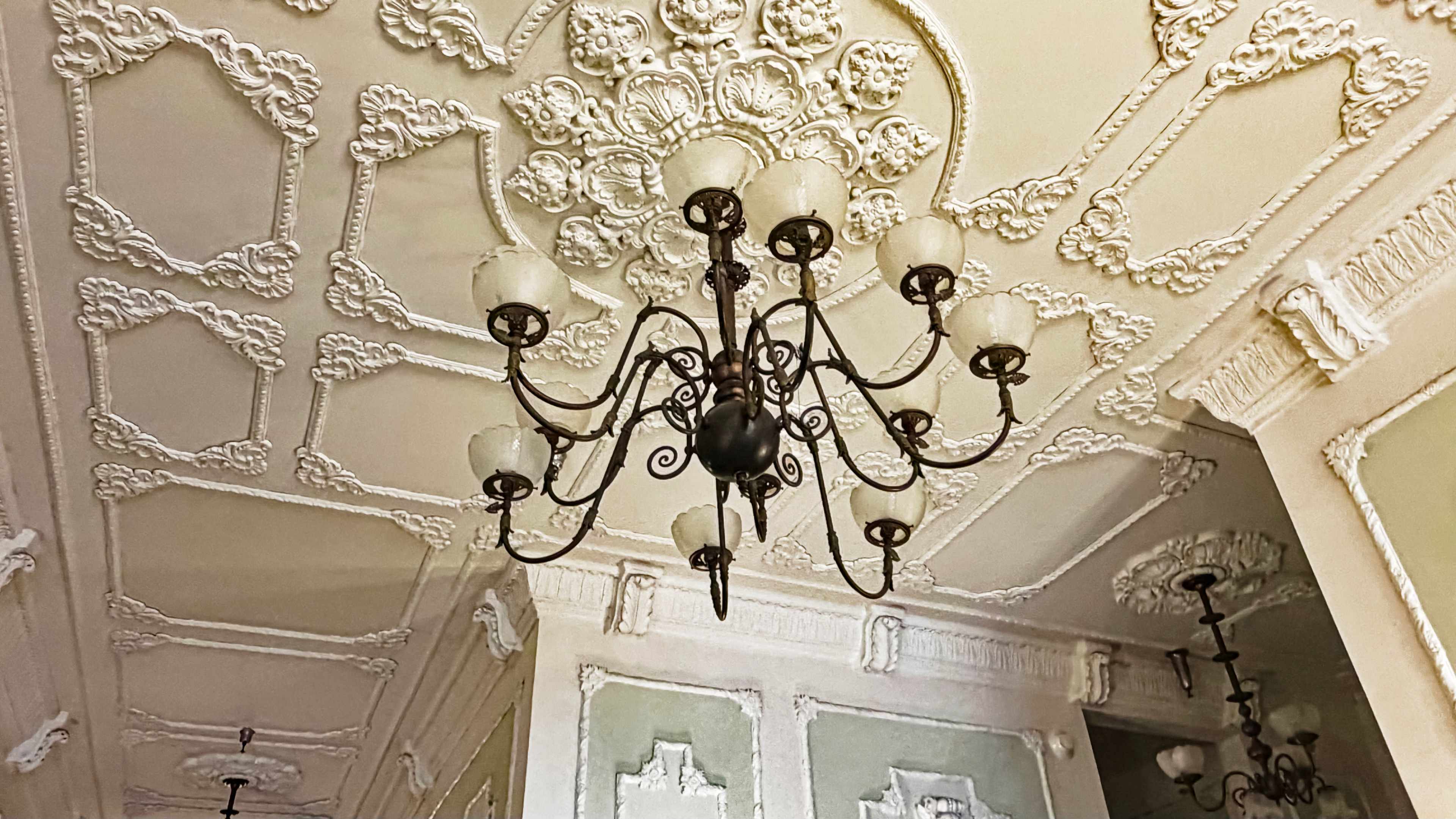 The image depicts an ornate ceiling with intricate moldings and a decorative chandelier.