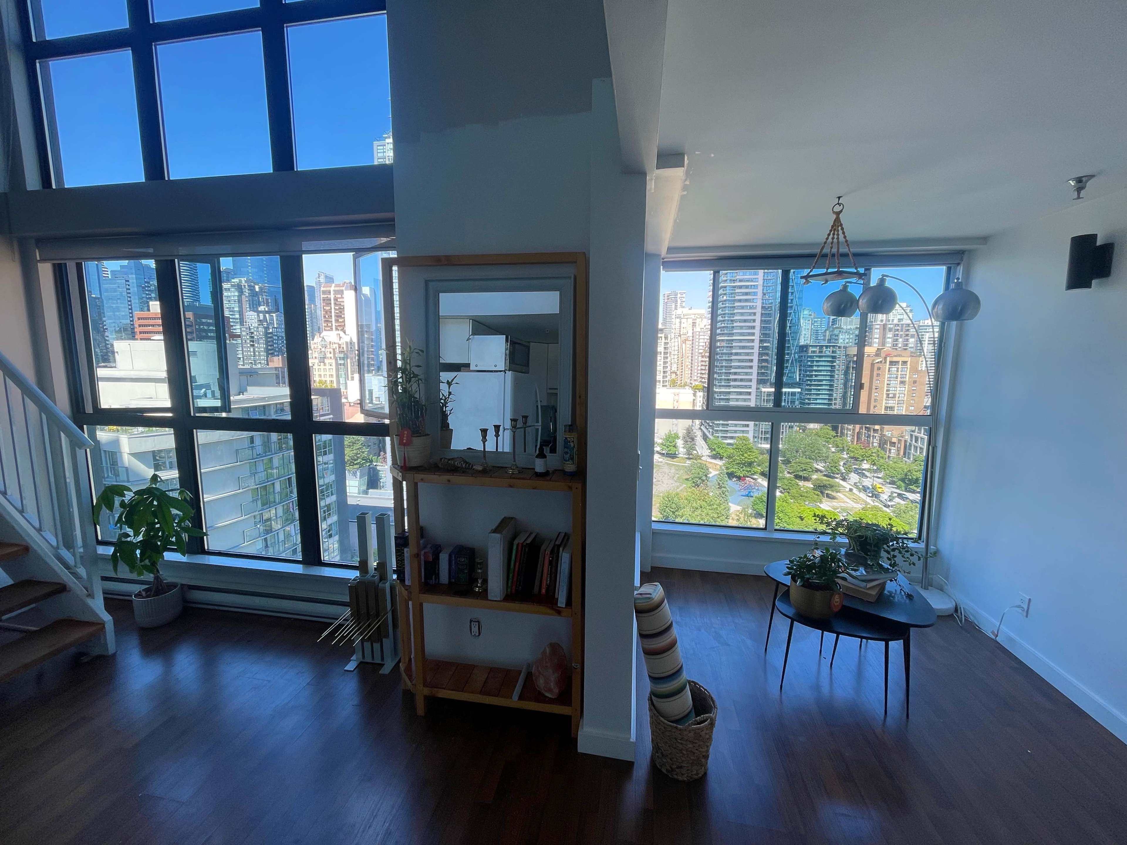 A bright, modern living space featuring large windows with a view of a cityscape, a wooden bookshelf, and a small round table.