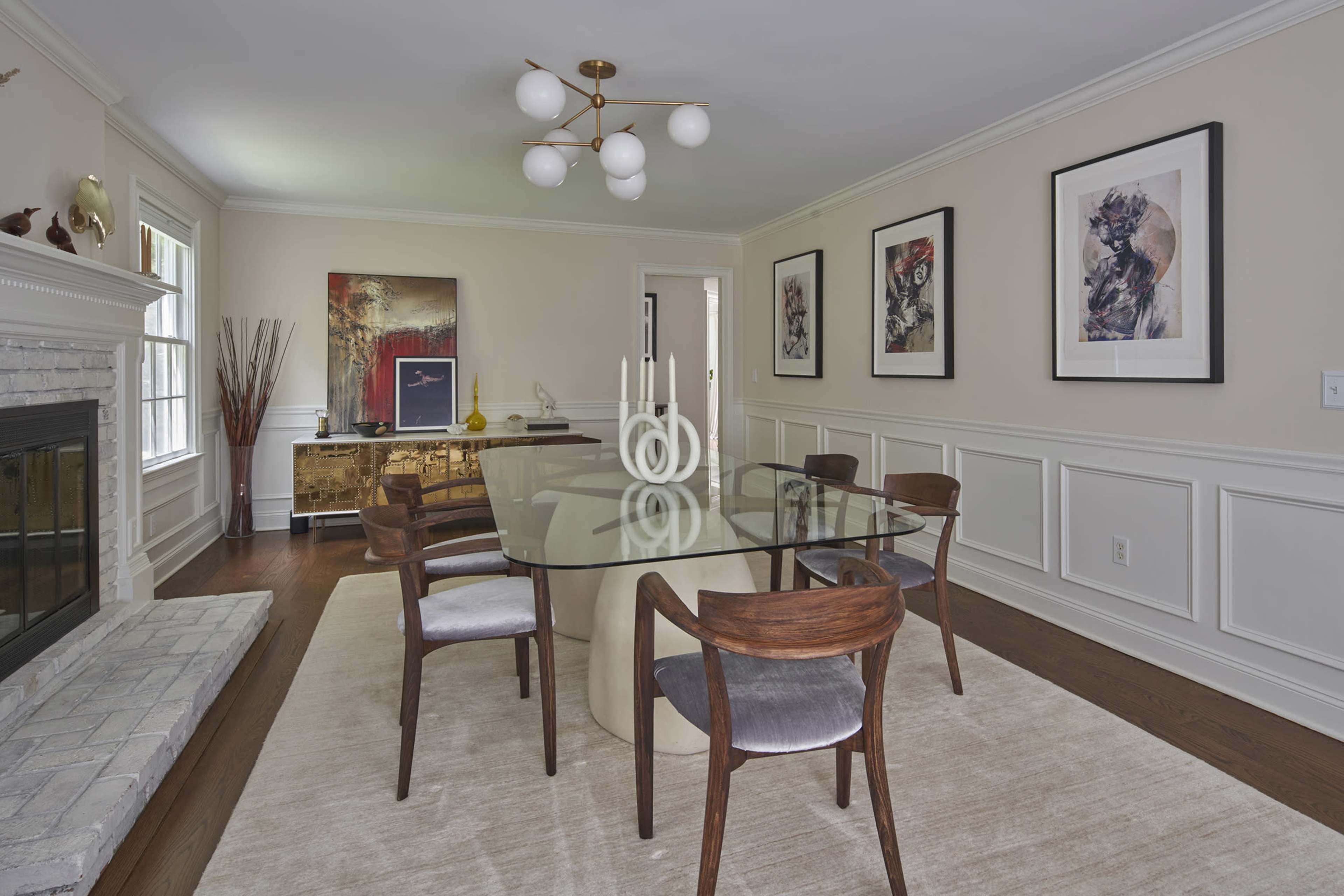 A modern dining room features a large round table surrounded by wooden chairs, with framed artwork on the walls and a fireplace to one side.