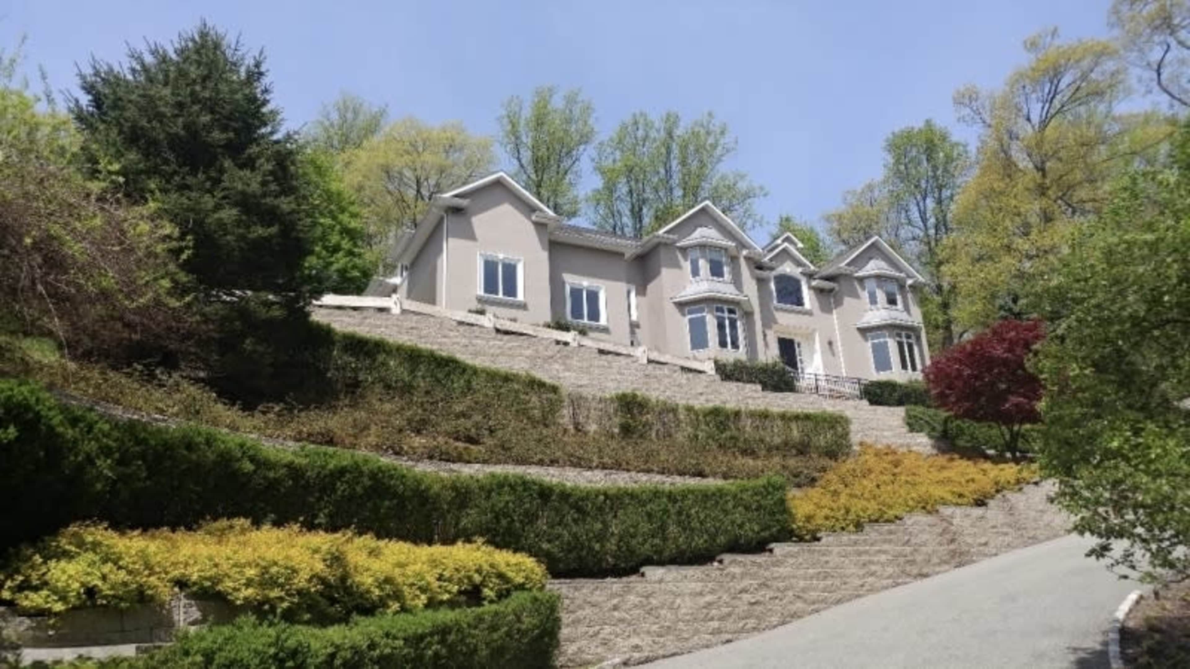 A large, multi-story house sits on a hillside, surrounded by neatly landscaped greenery and colorful shrubs.