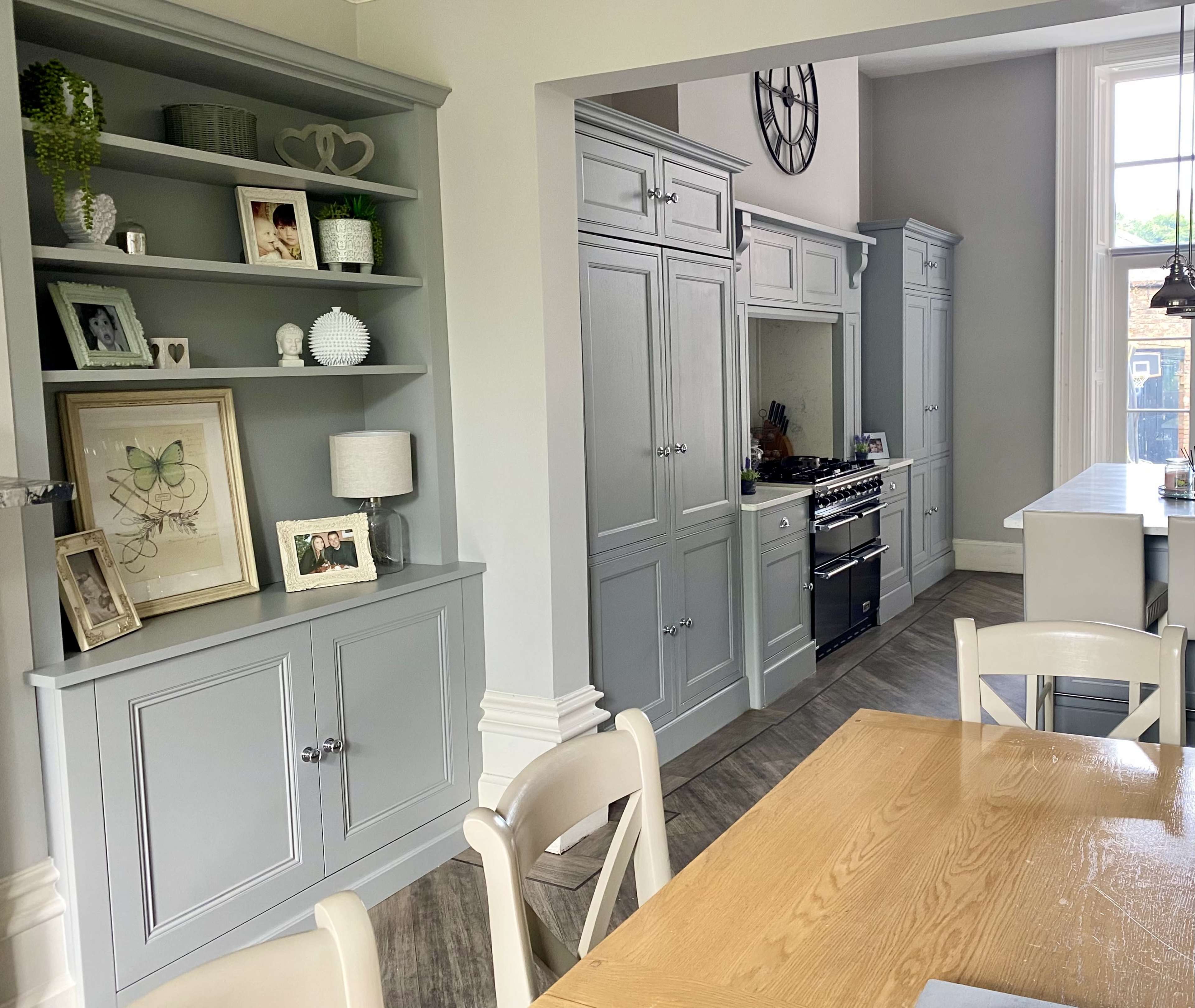 A modern kitchen and dining area with grey cabinetry, a wooden dining table, and a large clock on the wall.