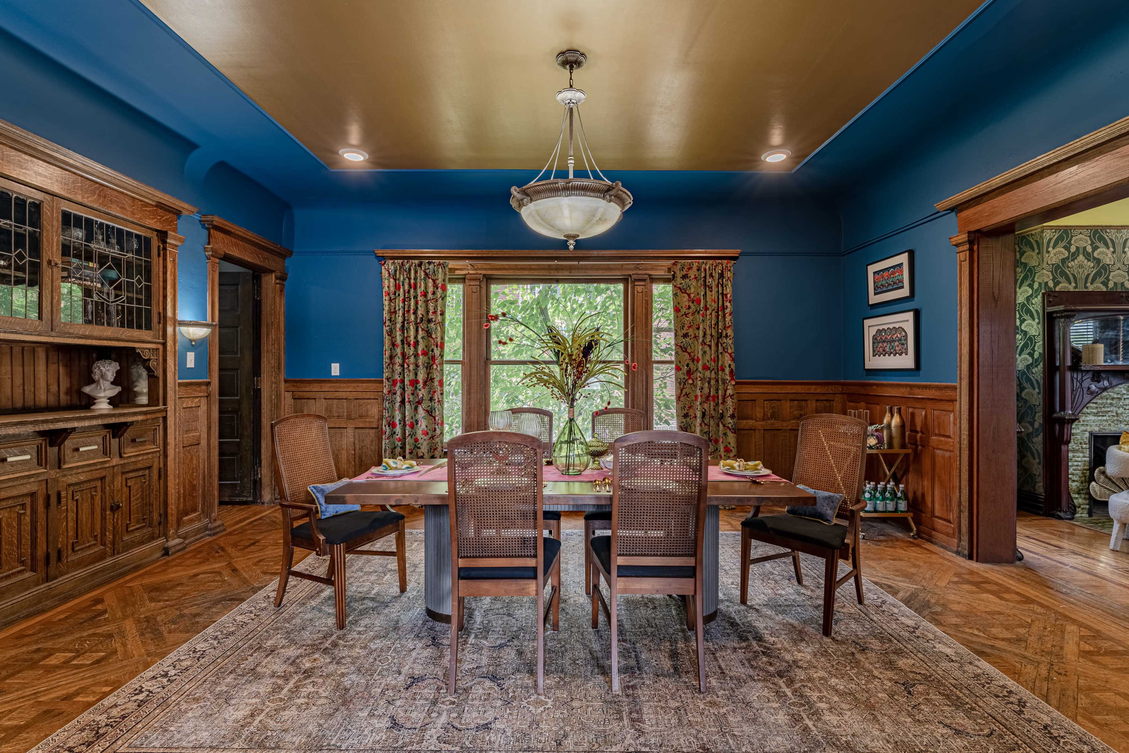 A dining room features a wooden table set for a meal, surrounded by four chairs, with decorative curtains, a chandelier, and richly stained wood paneling.