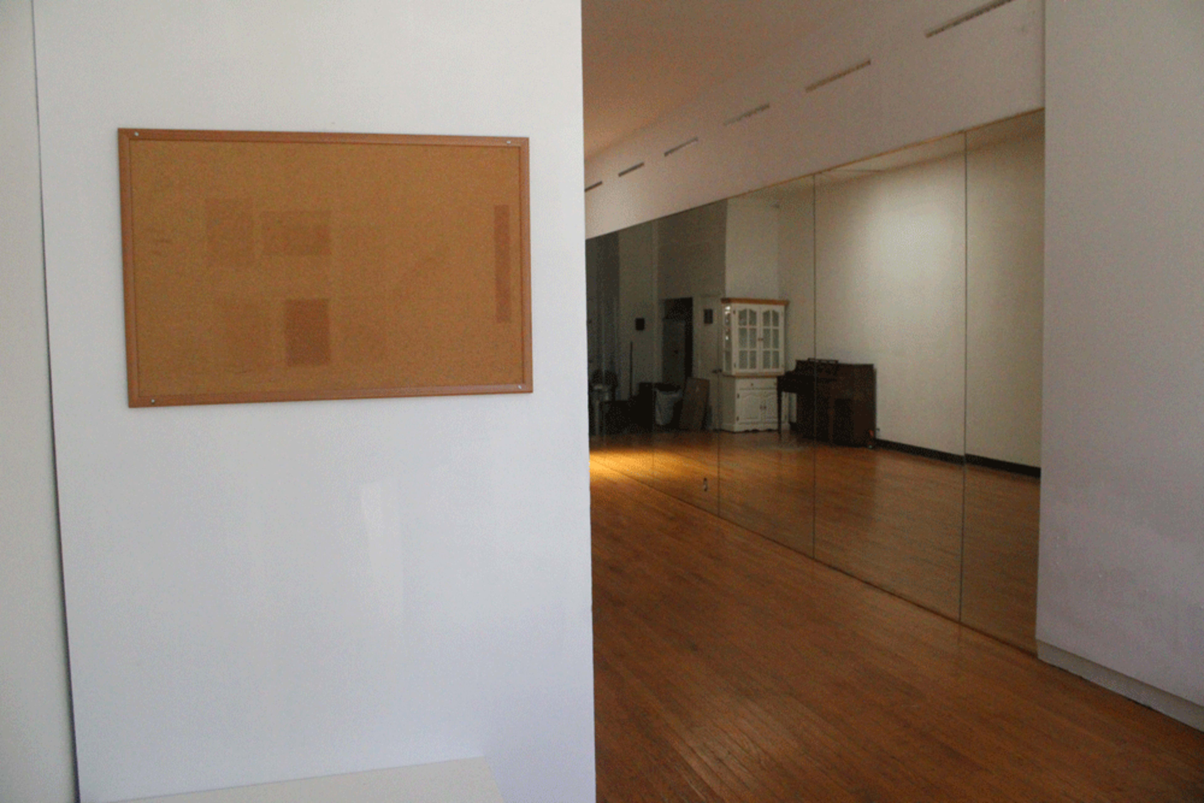 The image shows a spacious room with wooden flooring, featuring a wall-mounted corkboard and a large mirror on the opposite wall.