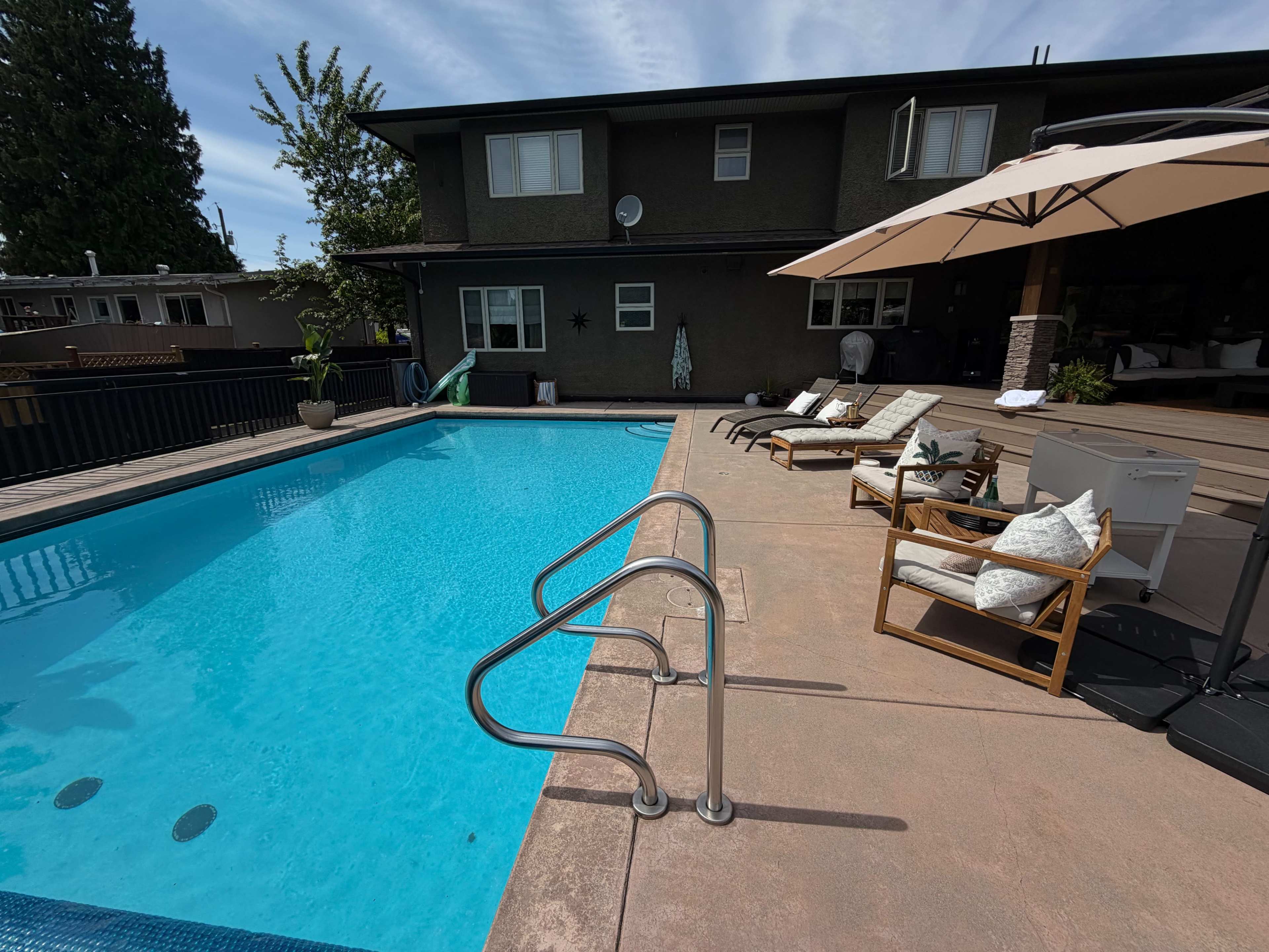 The image shows a swimming pool surrounded by lounge chairs and a large umbrella, beside a house with a wooden deck and greenery.