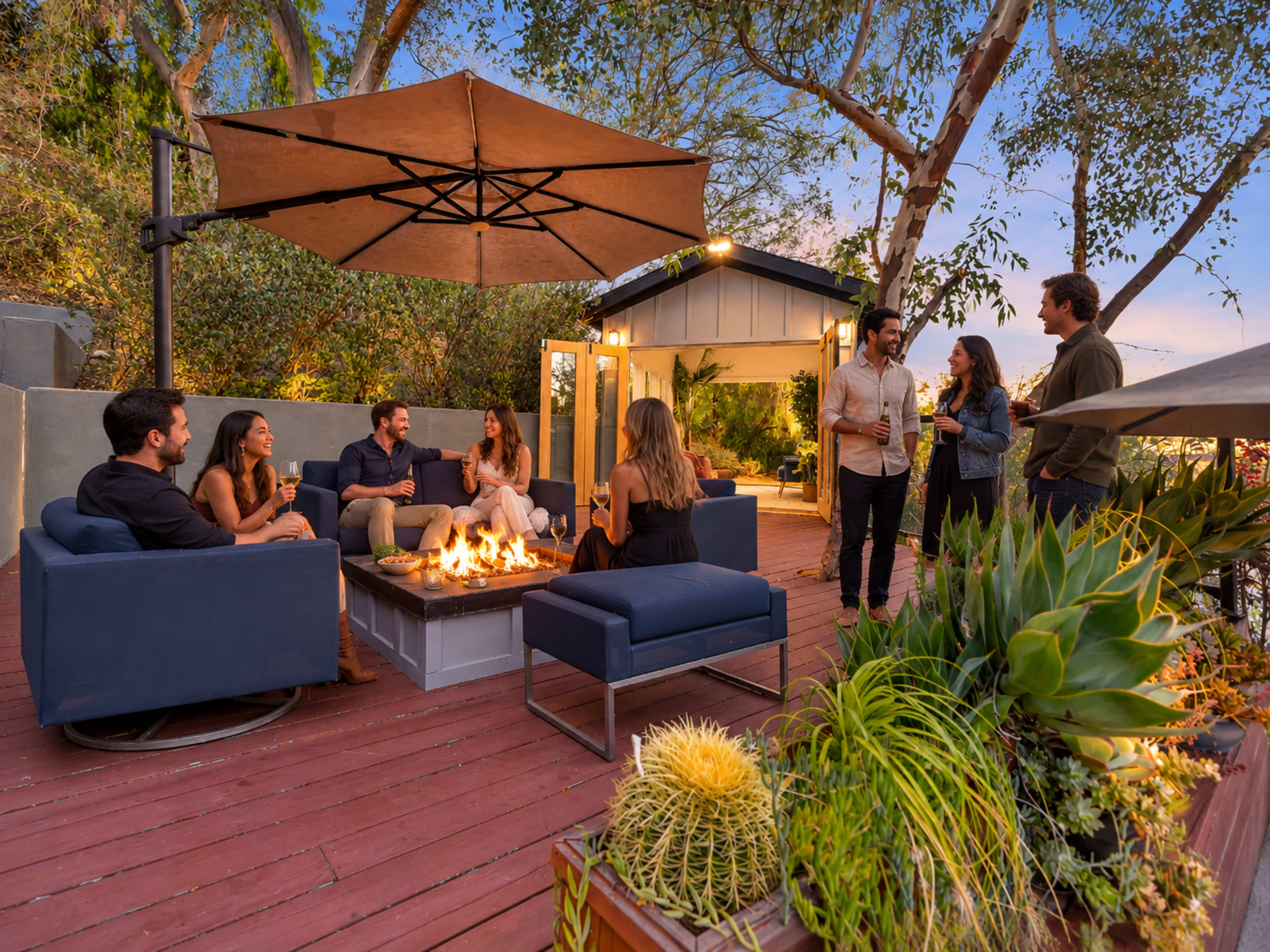 A group of people gathers around a fire pit on a wooden deck, socializing under a large umbrella in a scenic outdoor setting.