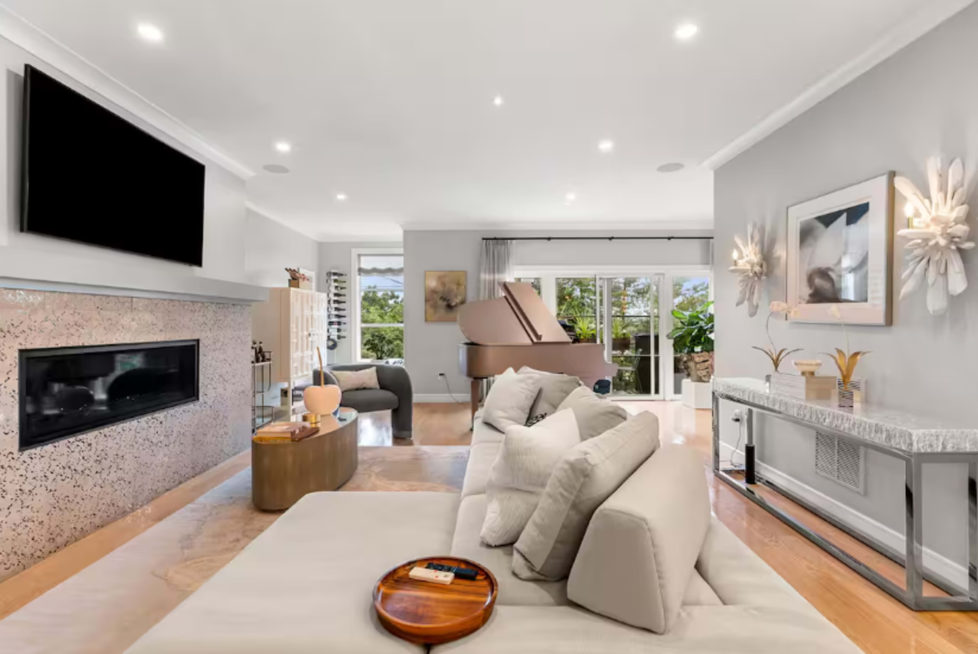 The image shows a modern living room featuring a large TV above a fireplace, a beige sofa, a coffee table, and a grand piano in the background, with natural light coming through large windows.