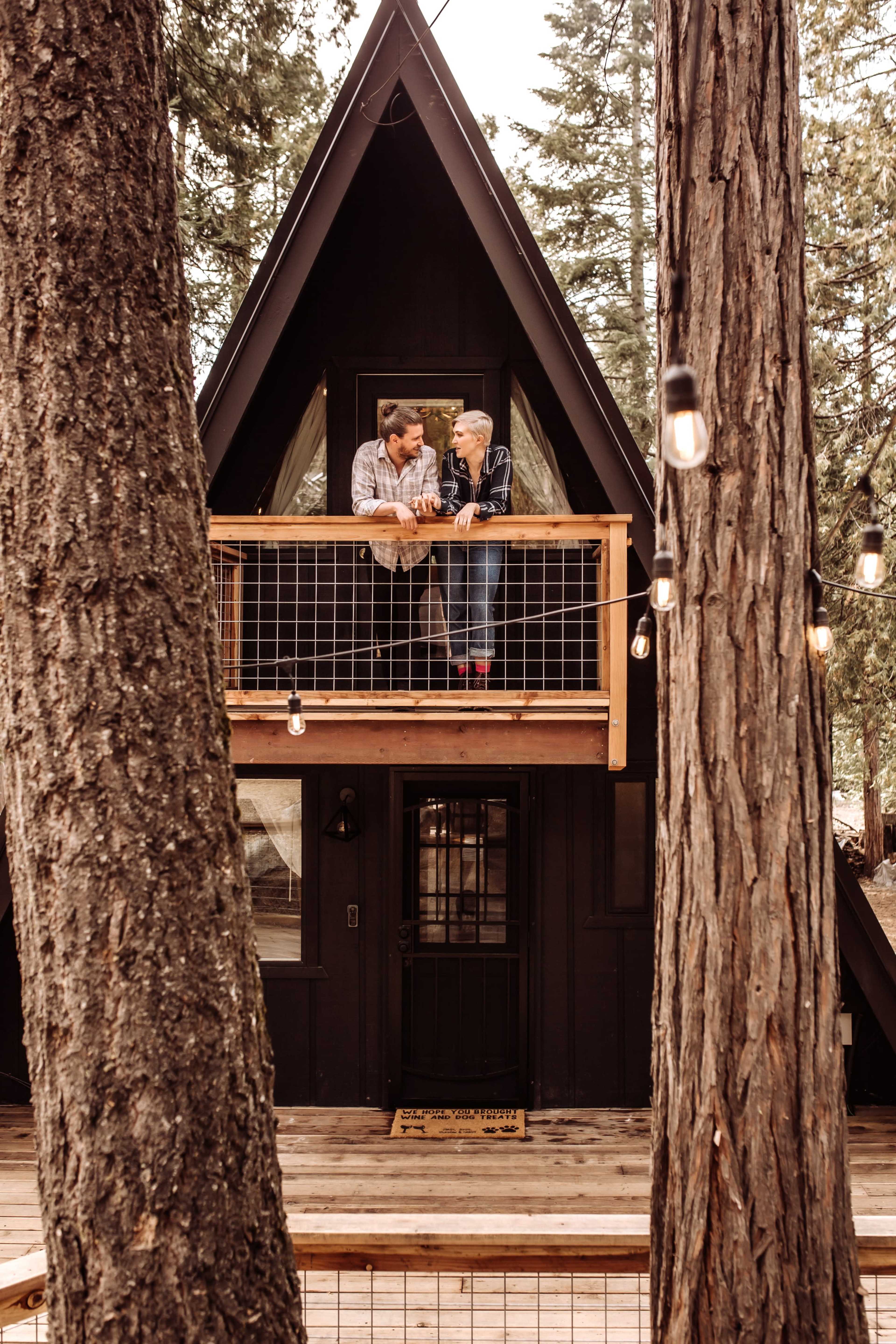 A couple stands on the balcony of an A-frame cabin surrounded by tall trees.