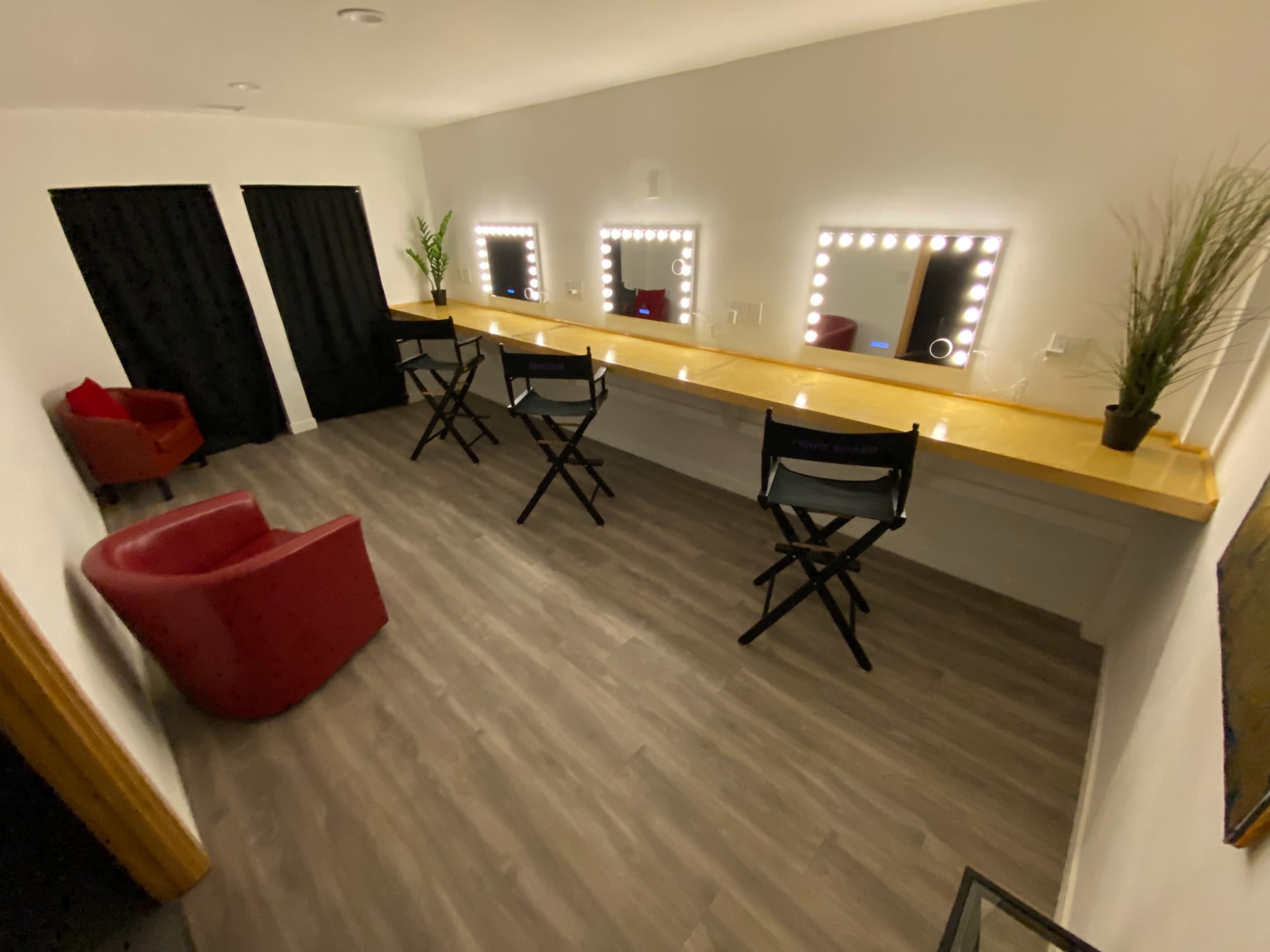A dressing room with a long mirror-lit vanity, two director's chairs, a red armchair, and black curtains in the background.