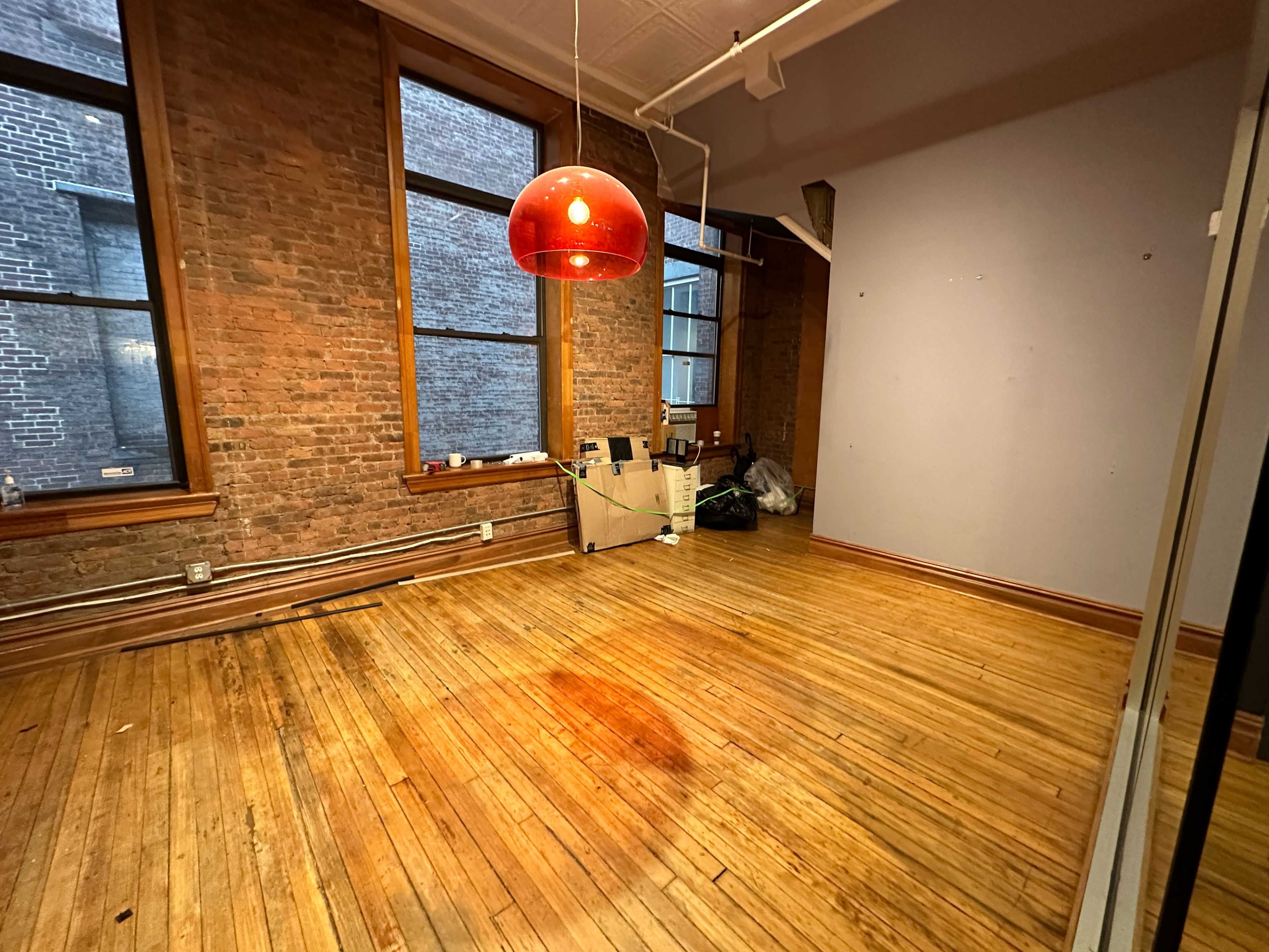 A vacant room with exposed brick walls, wooden floors, large windows, and a red pendant light.