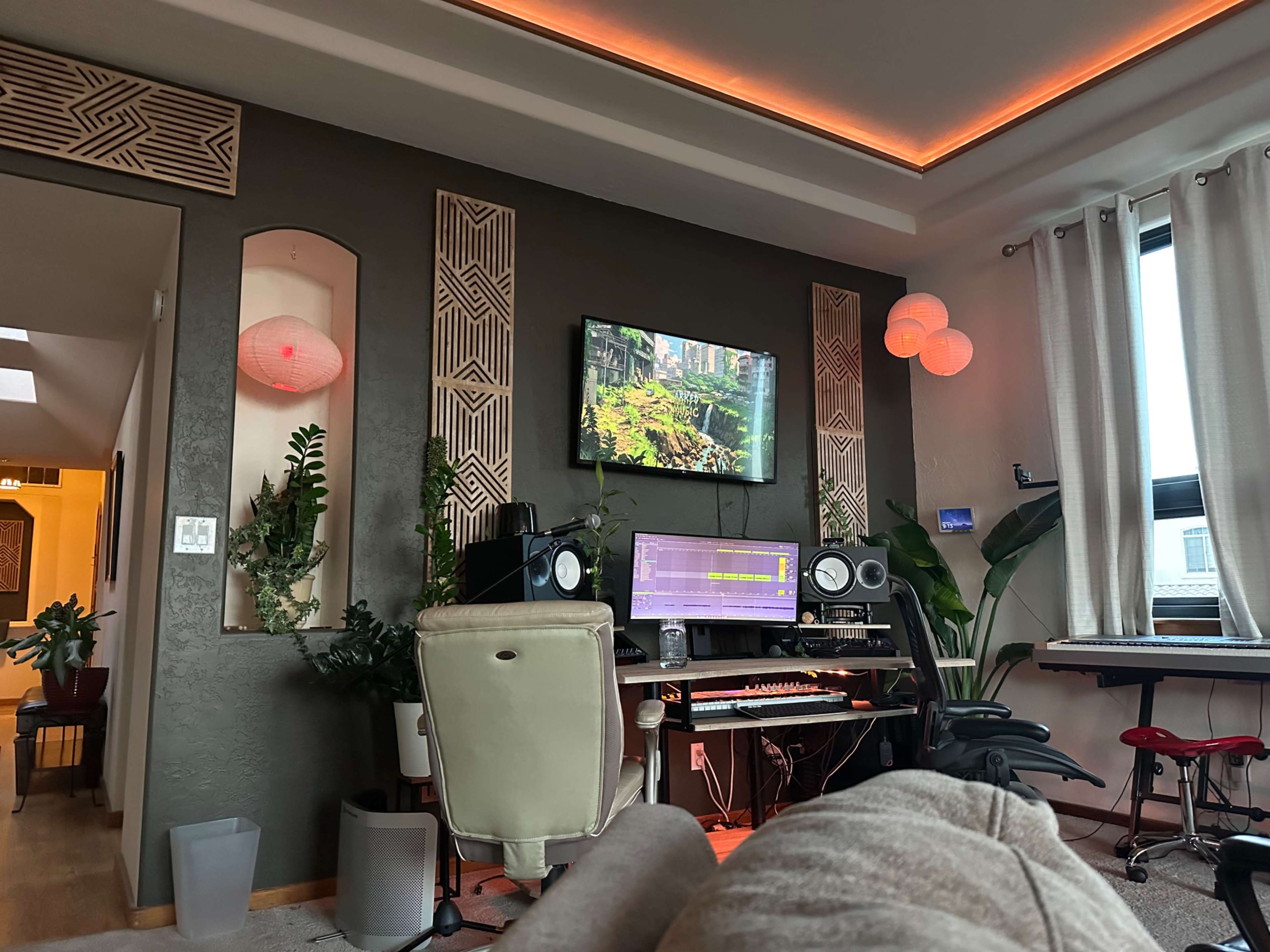 The image depicts a home music studio featuring a computer workstation with audio equipment, a large monitor displaying a nature scene, and plants along the walls, illuminated by ambient lighting.