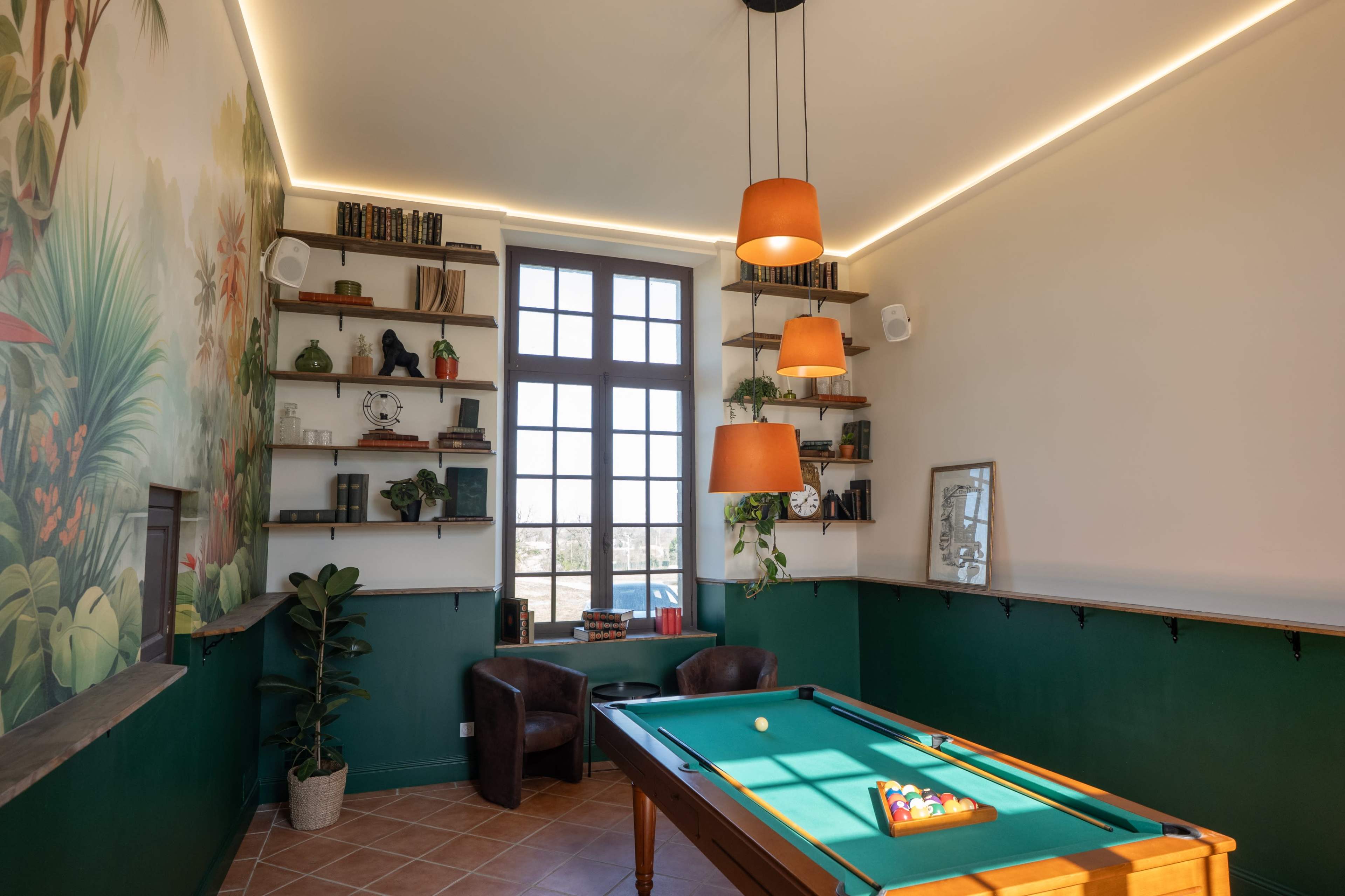 A pool table is positioned in the center of a room with green walls, decorated shelves, and large windows letting in natural light.