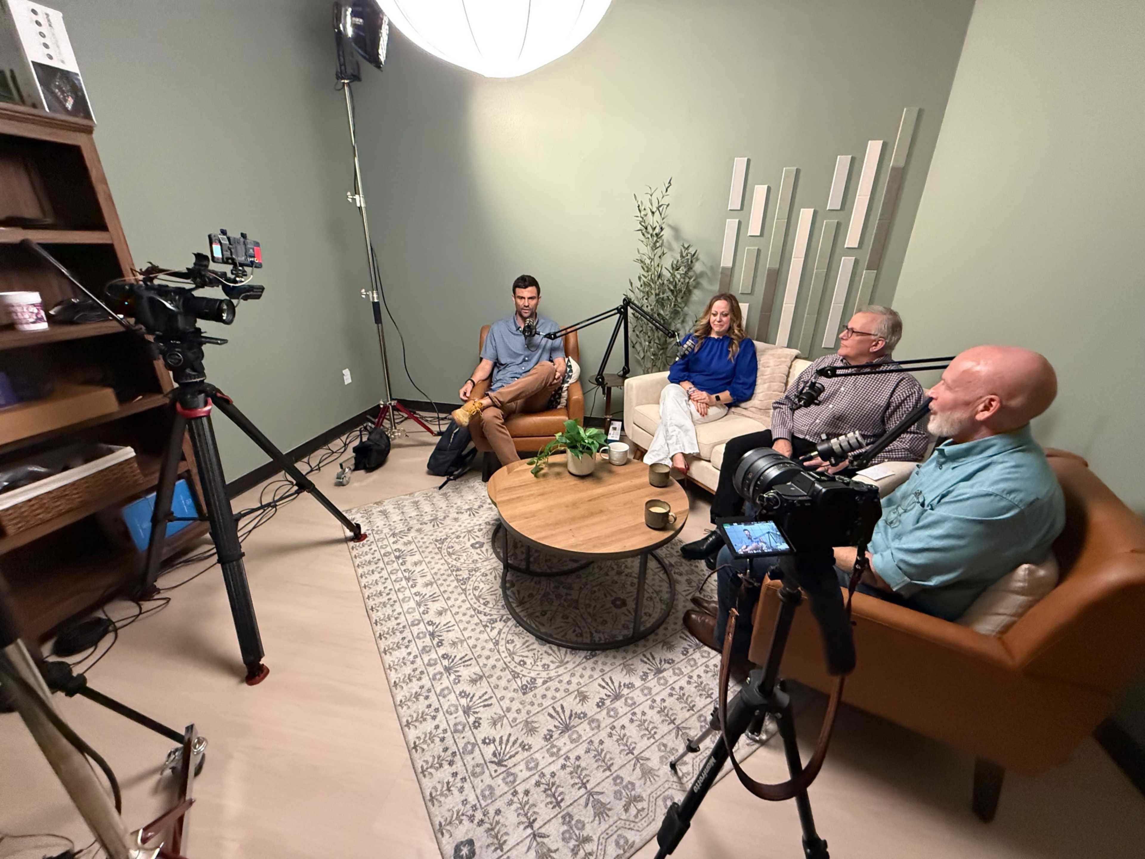 A group of four people is seated on a couch in a recording studio, surrounded by video equipment and studio lights.