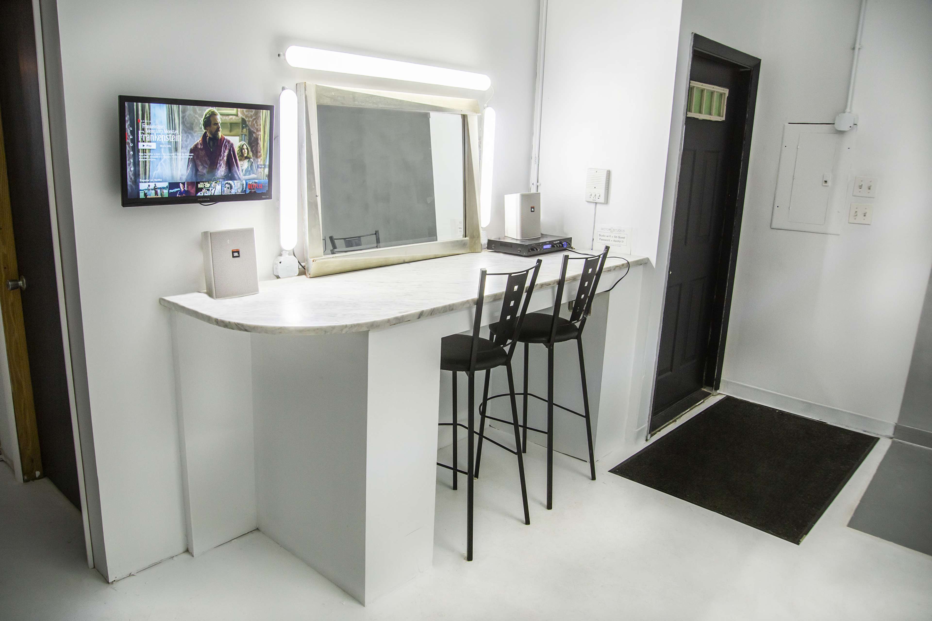 A minimalist vanity area with a marble countertop, two black bar stools, a wall-mounted mirror, and a television display.
