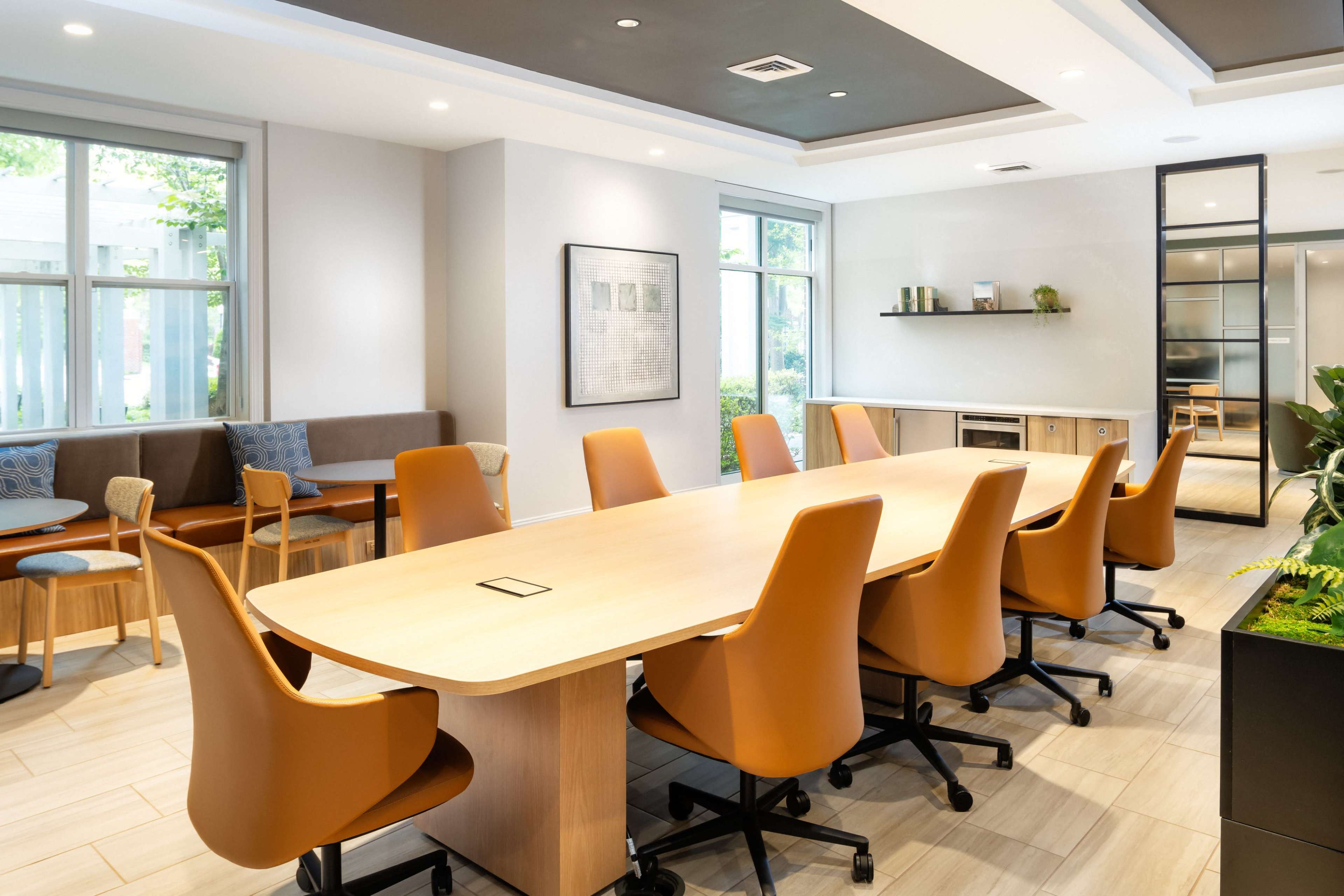 The image shows a modern meeting room with a long wooden table surrounded by orange chairs and large windows allowing natural light to enter.