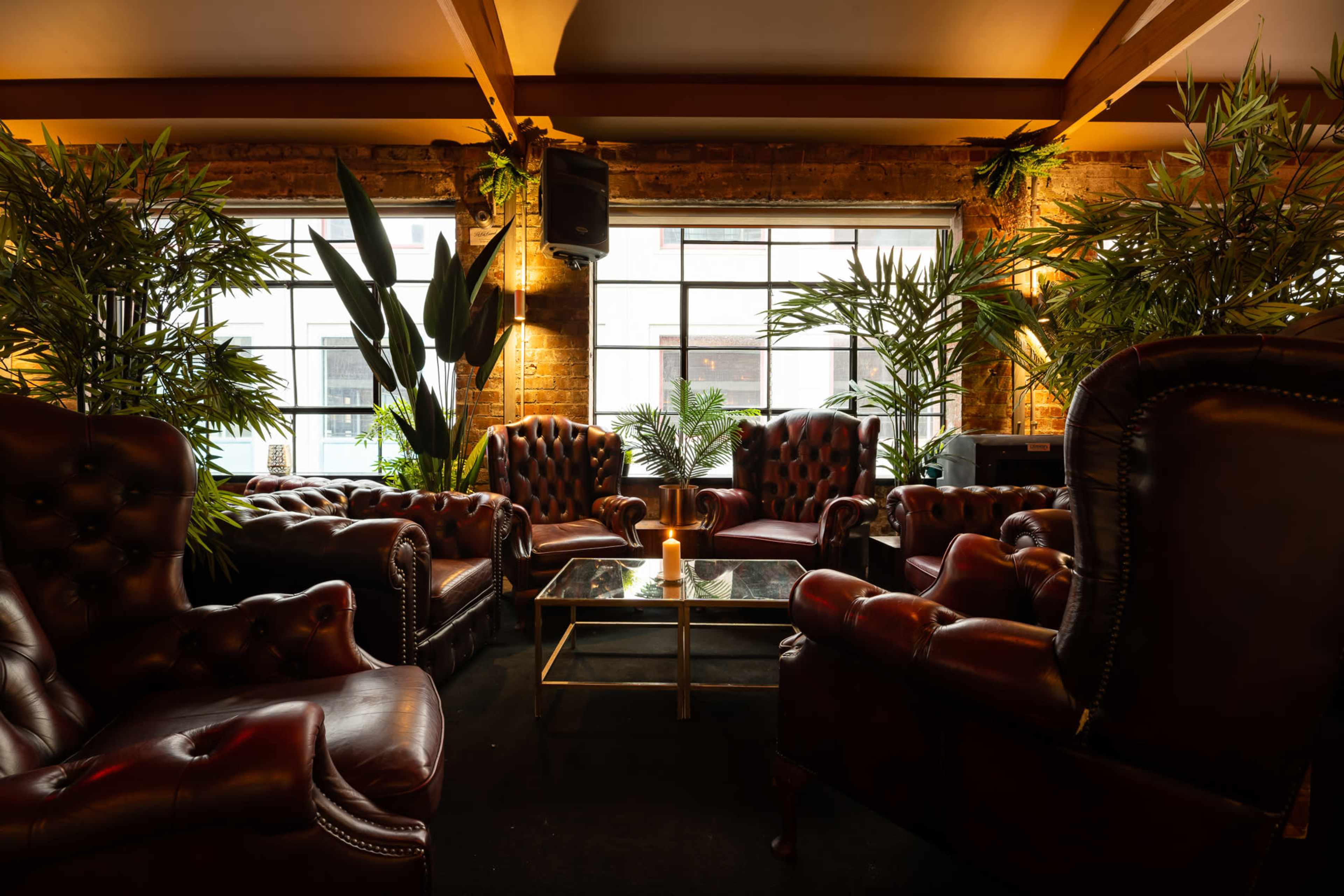 A cozy seating area features leather armchairs arranged around a coffee table, surrounded by potted plants and large windows.