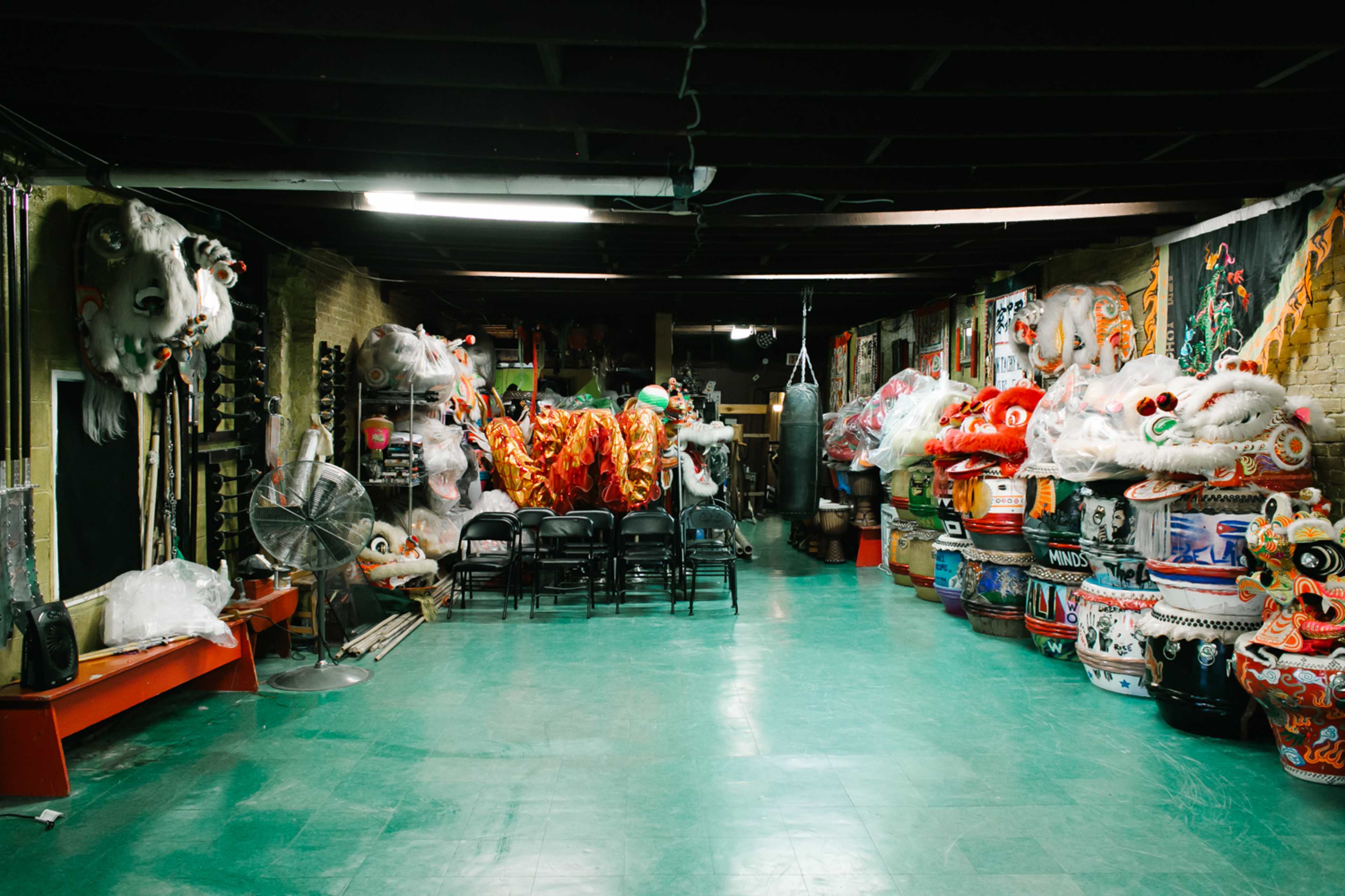 A dimly lit room filled with vibrant lion dance costumes and props, with black chairs arranged in the center.
