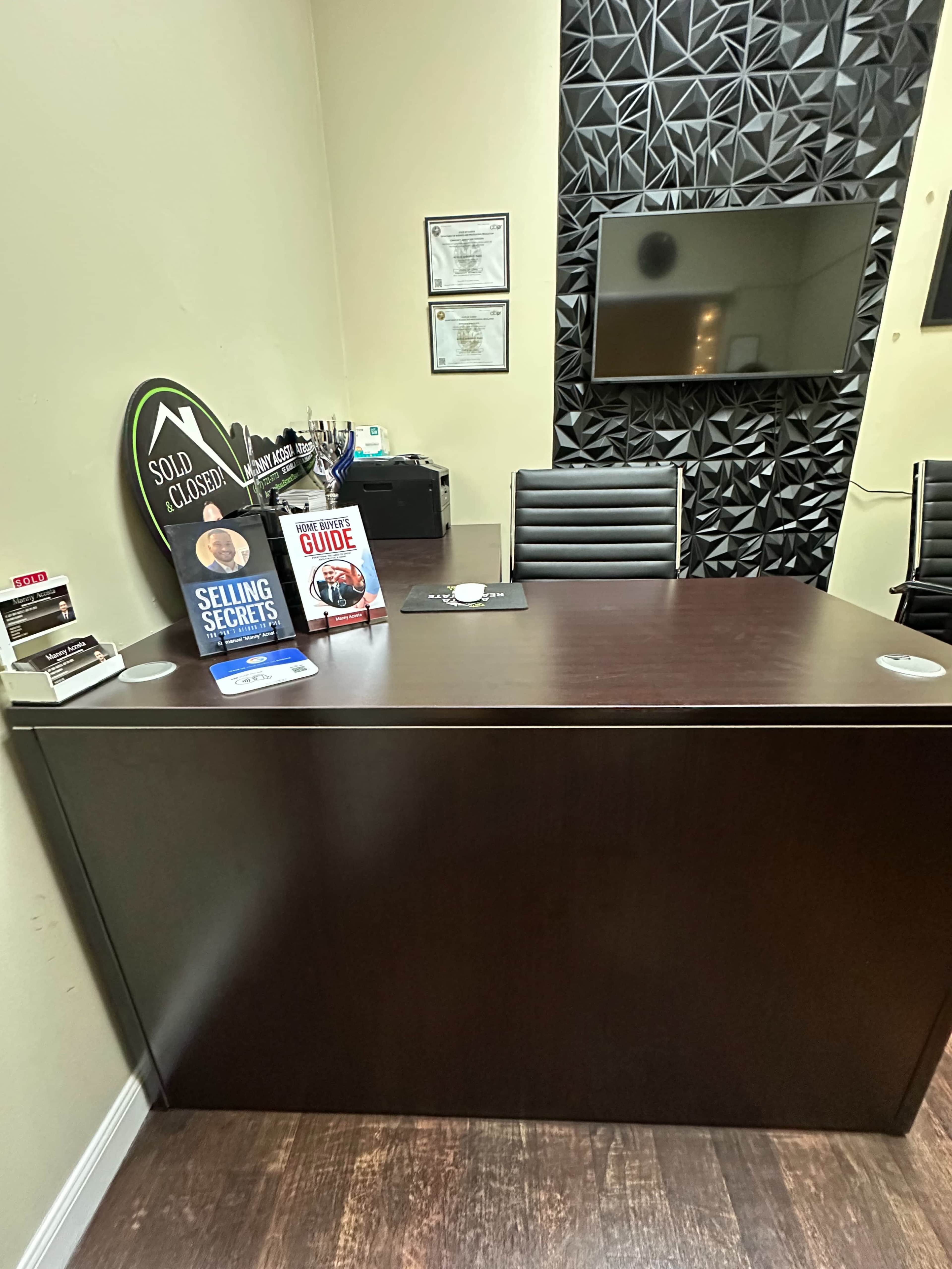 The image shows a desk in an office with a black geometric accent wall, featuring books about sales and a television mounted on the wall.