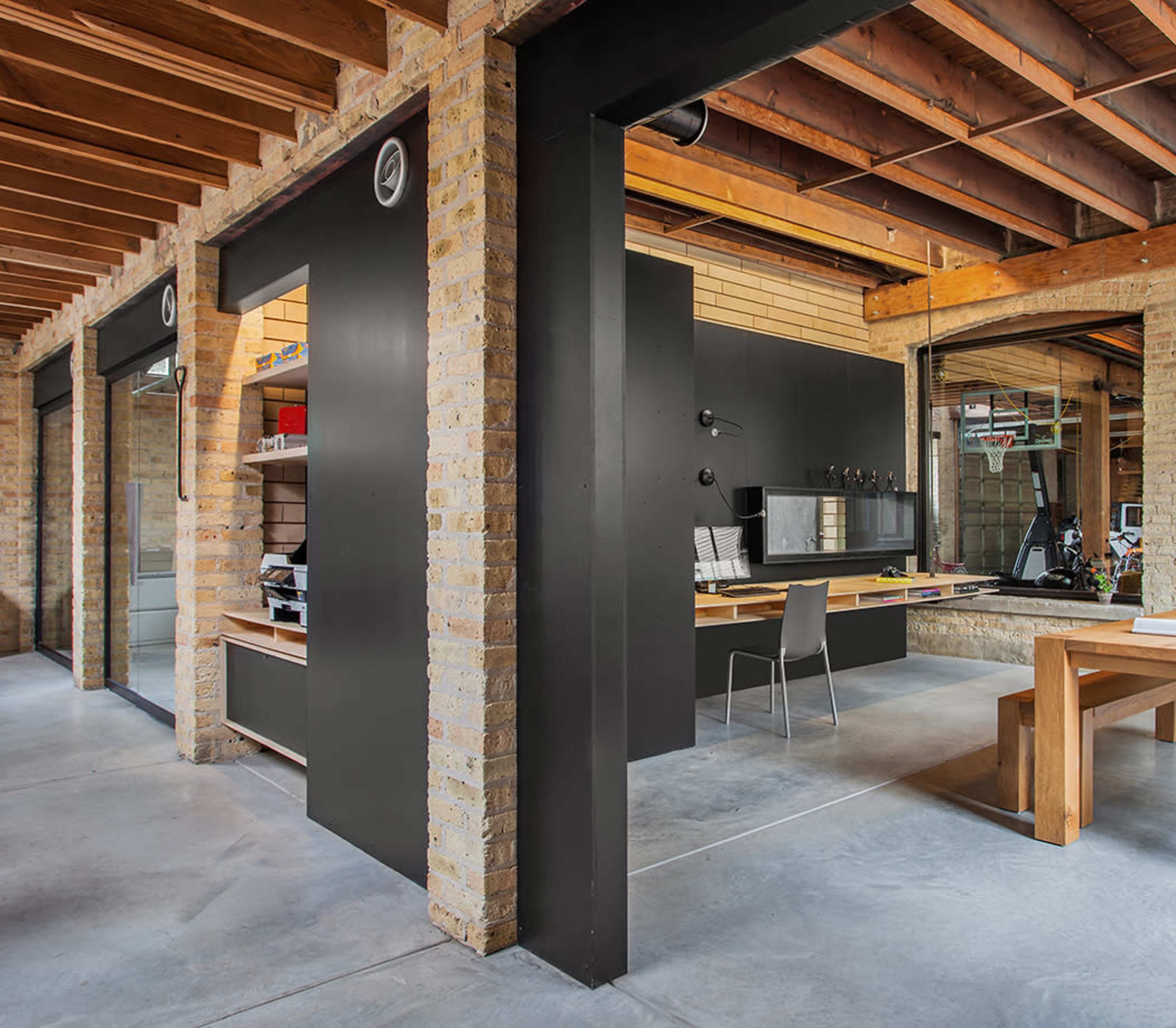 The image shows a modern office space featuring exposed wooden beams, brick walls, and minimalist black furniture, including a large desk and a simple chair.