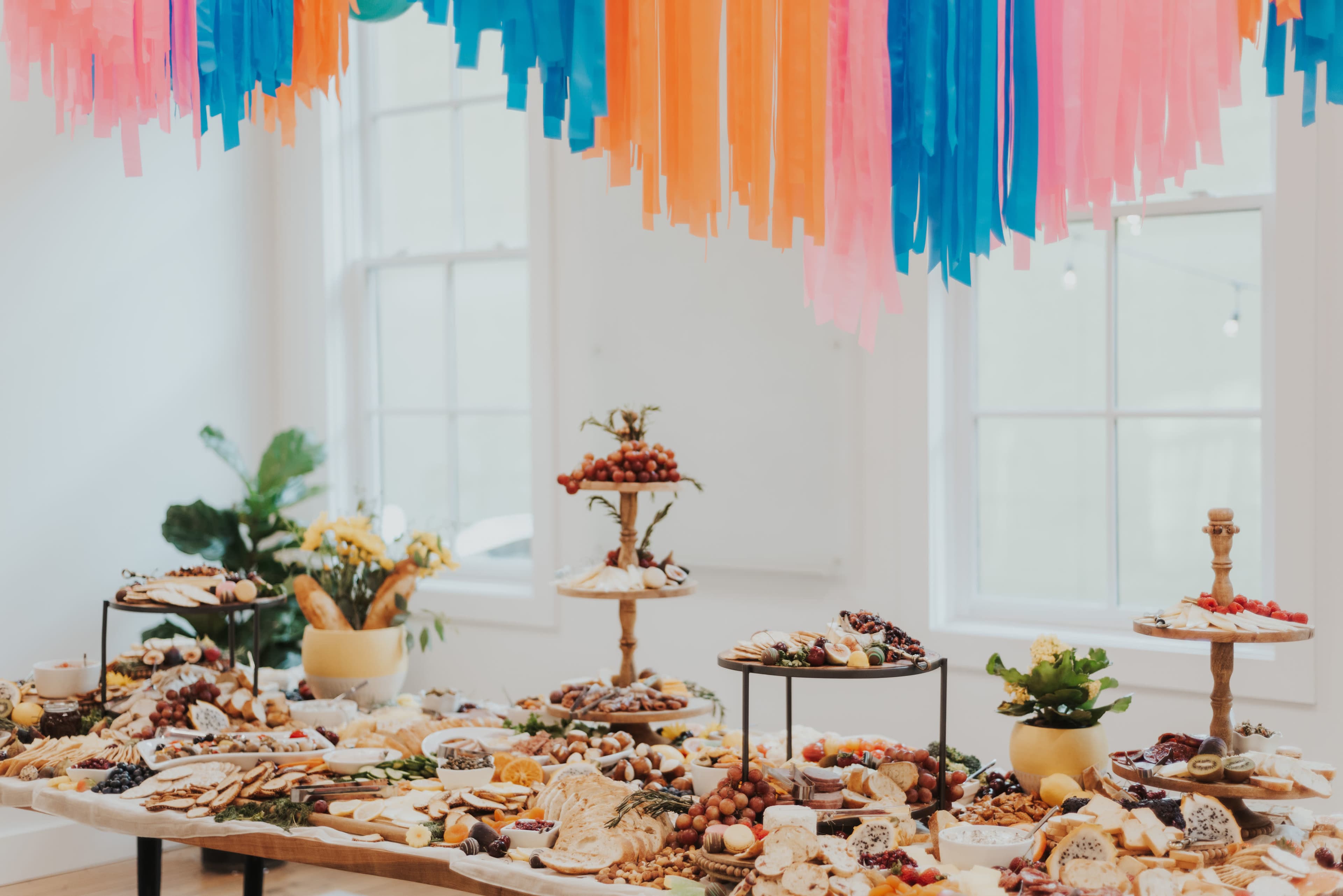 A large spread of assorted foods and snacks is elegantly displayed on tables beneath colorful streamers in a bright room.