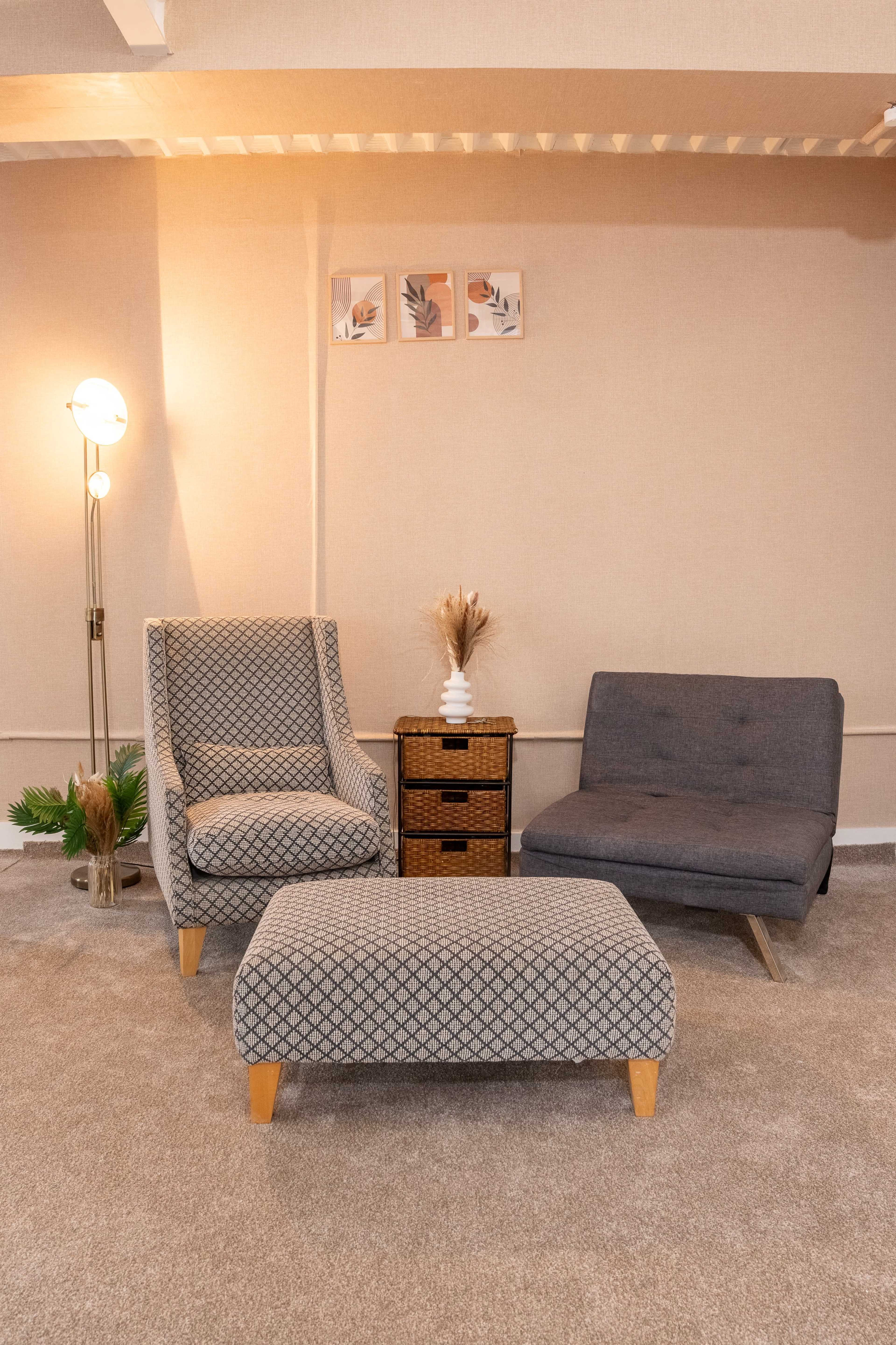 A cozy seating area features a patterned armchair, a grey loveseat, a patterned ottoman, and a wooden side table adorned with decorative elements.