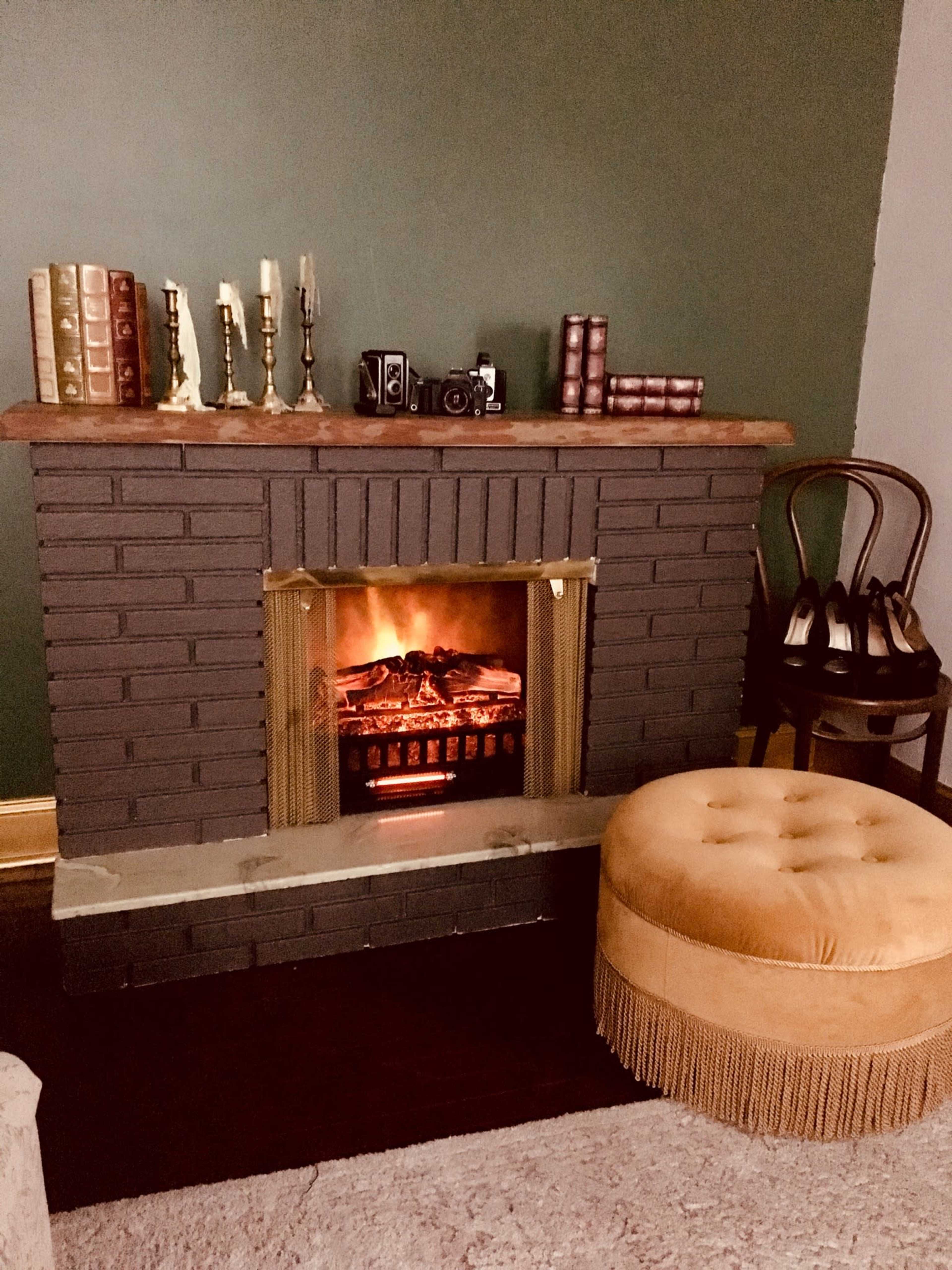 A vintage-style fireplace with an electric insert is flanked by candlesticks and old books, accompanied by a round tufted ottoman and a chair with shoes resting nearby.