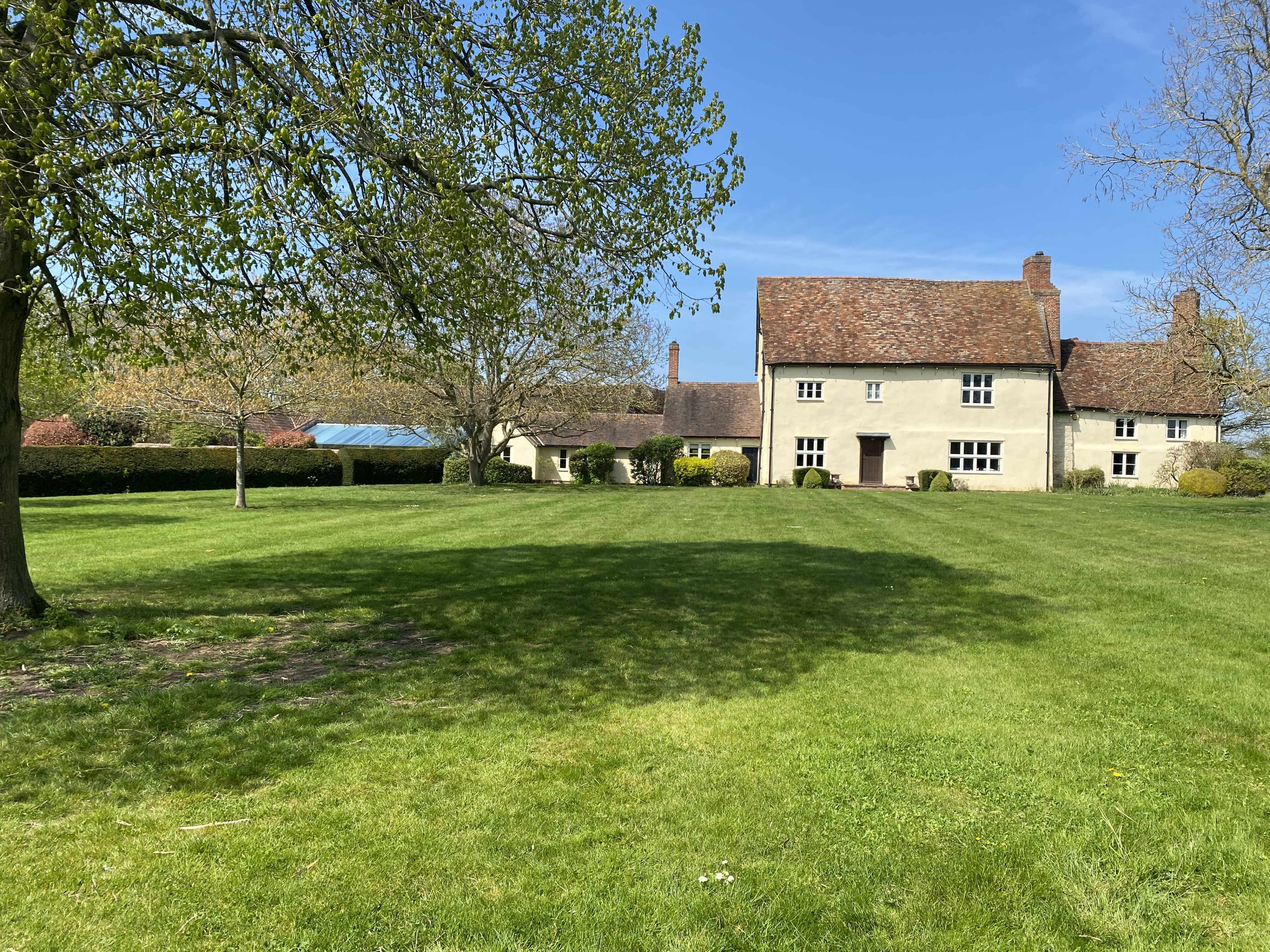 A two-story house with a brown, tiled roof is surrounded by a grassy lawn and trees under a clear blue sky.