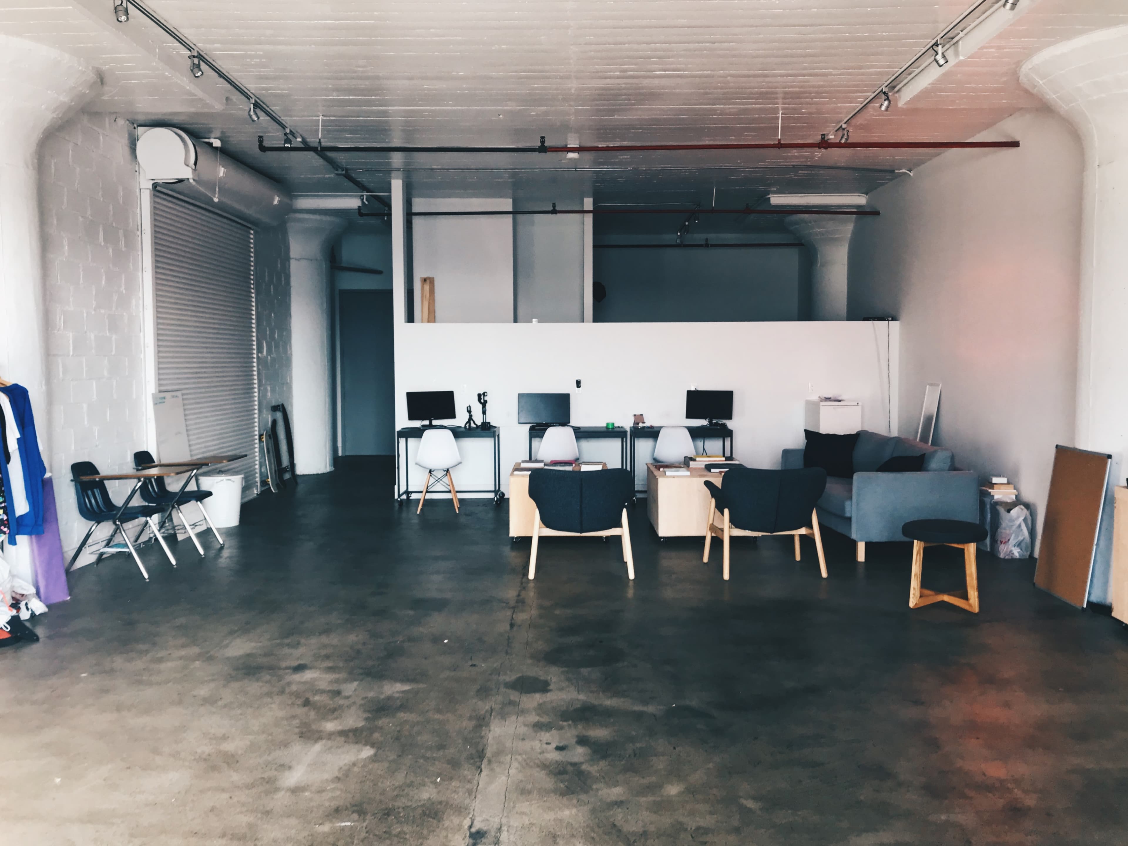 The image shows an empty workspace with several desks, chairs, and a small seating area in a well-lit, spacious setting.