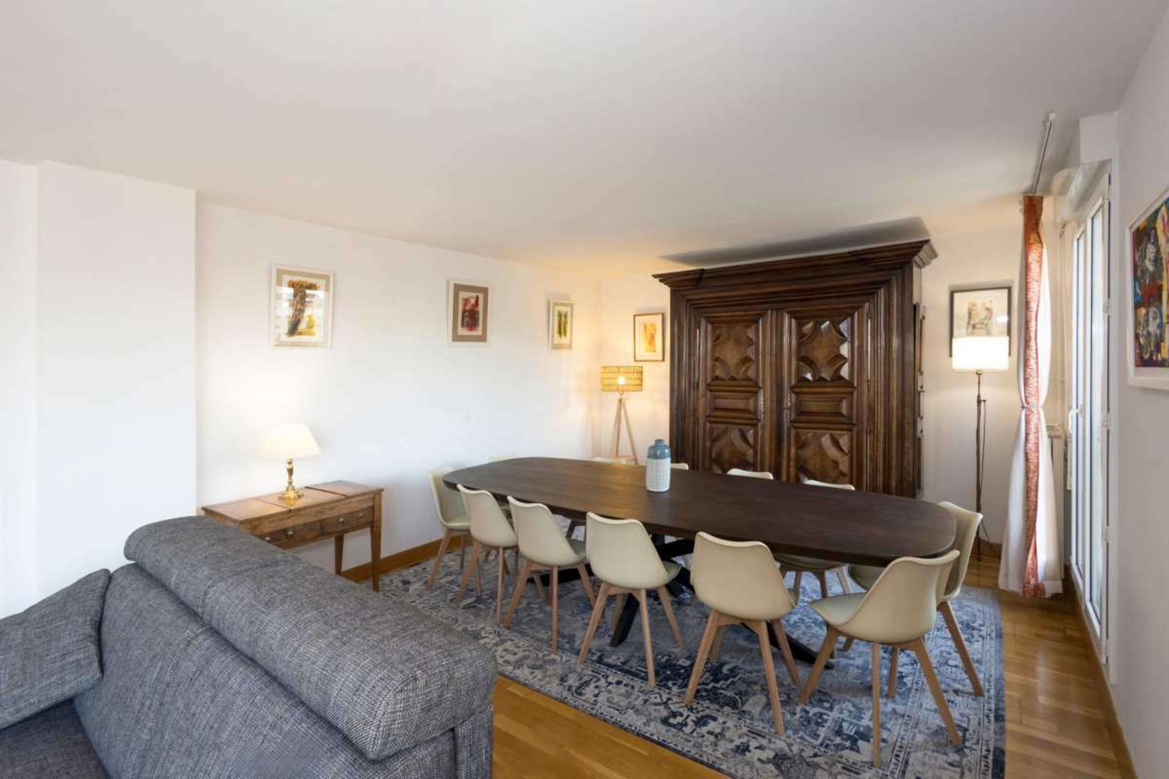 The image shows a modern dining room with a large wooden table, eight light-colored chairs, a decorative cabinet, and ambient lighting.