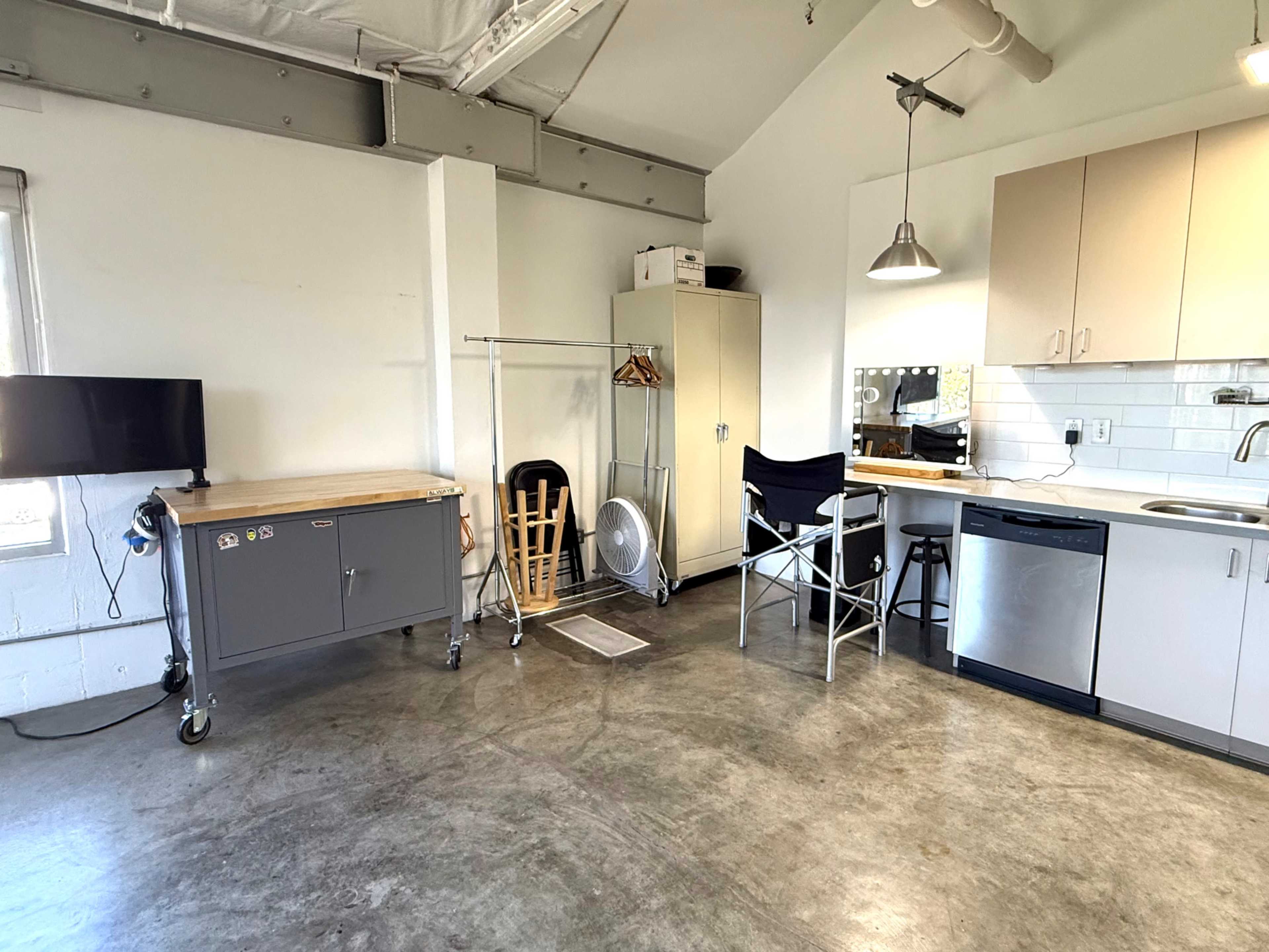 The image shows a small apartment interior with a kitchenette, a folding chair, a rolling kitchen cart, and a television mounted on the wall.