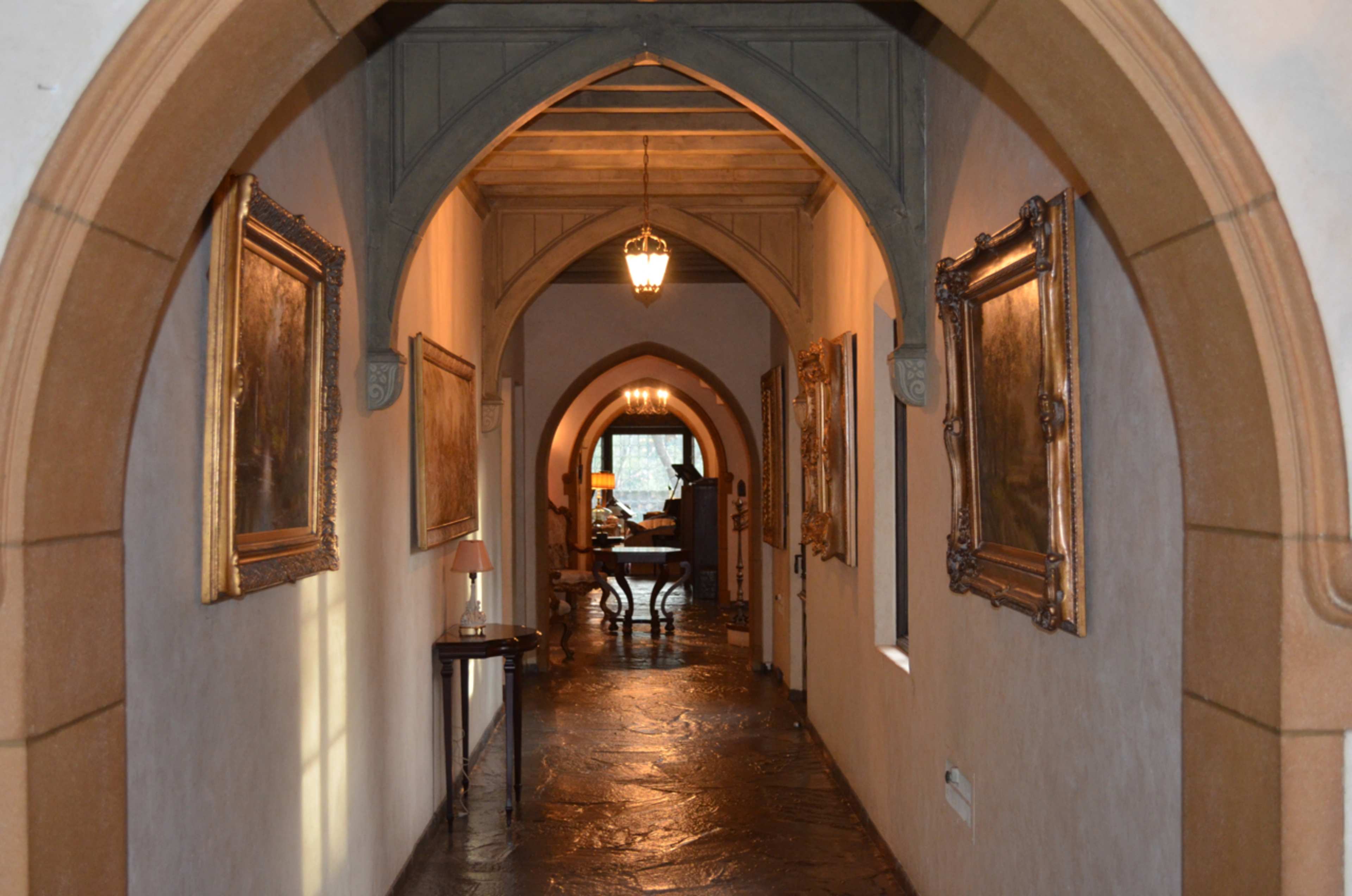 A long, arched hallway with stone flooring and framed paintings on the walls leads to a warmly lit room at the end.