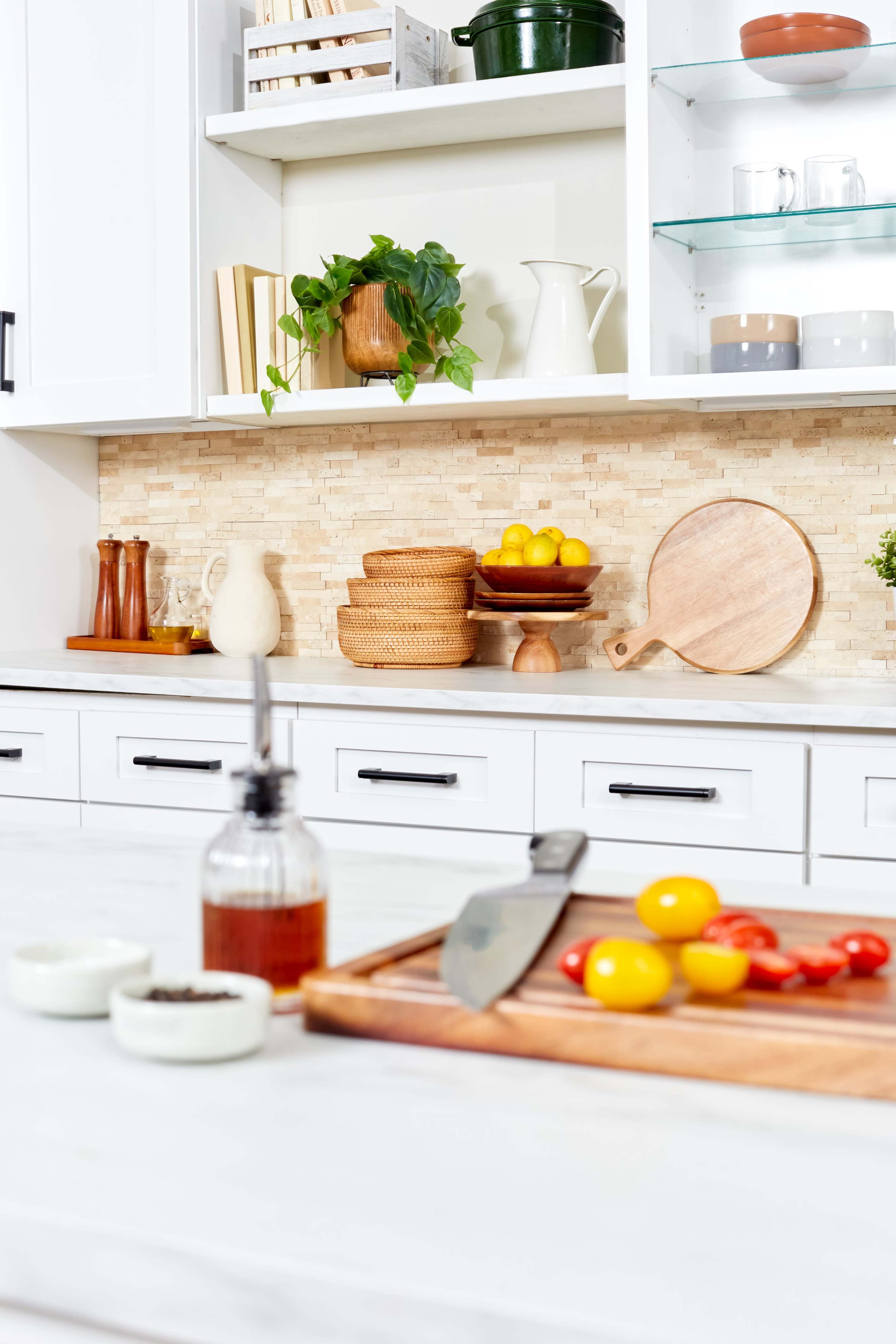 A modern kitchen with a marble countertop, featuring a wooden cutting board with sliced lemons and tomatoes, and organized shelves with dishes and decorative items.