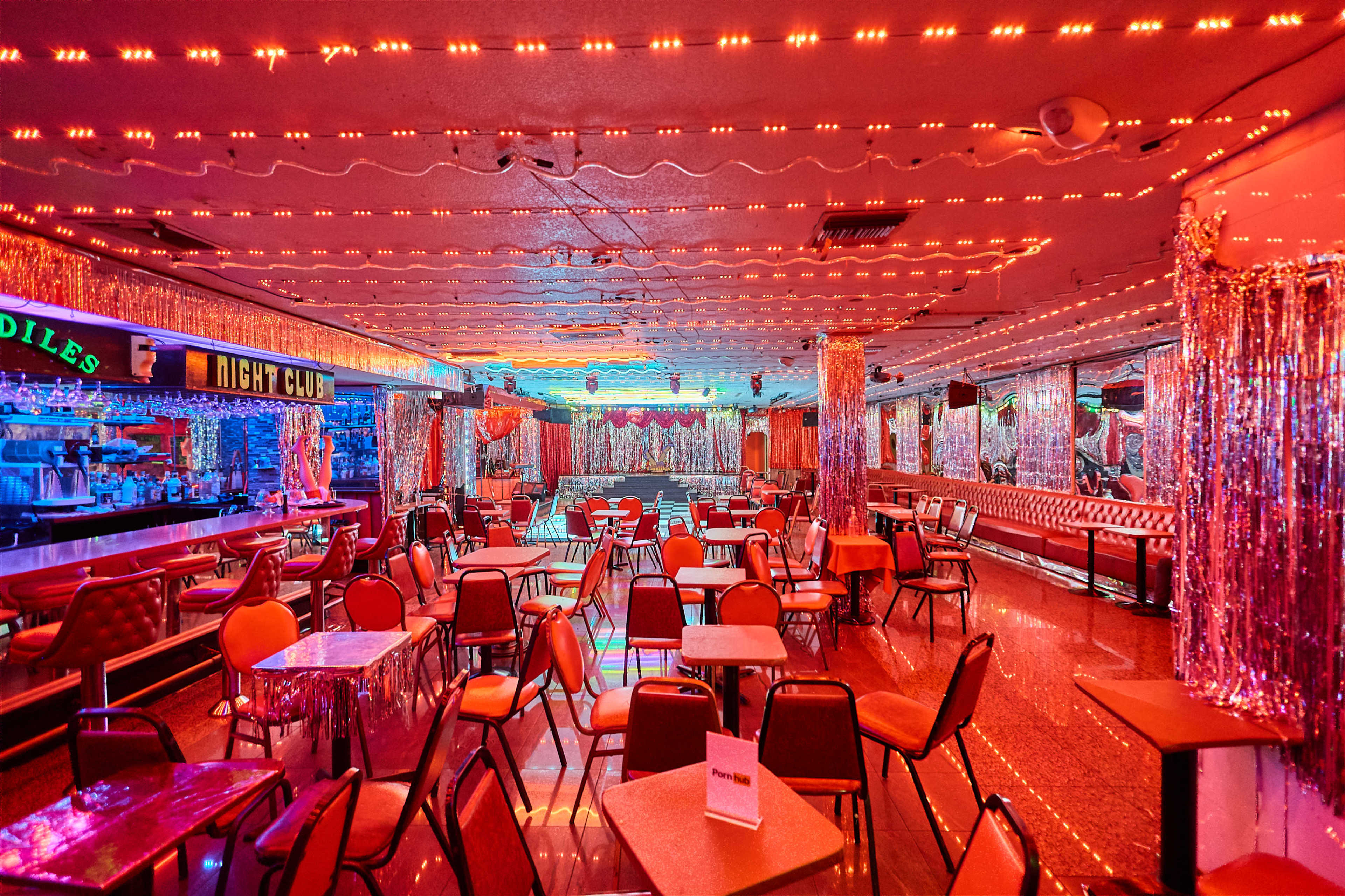 The image shows an empty night club interior adorned with red and blue lights, tables, and chairs arranged in rows, with sparkling decorations on the walls.