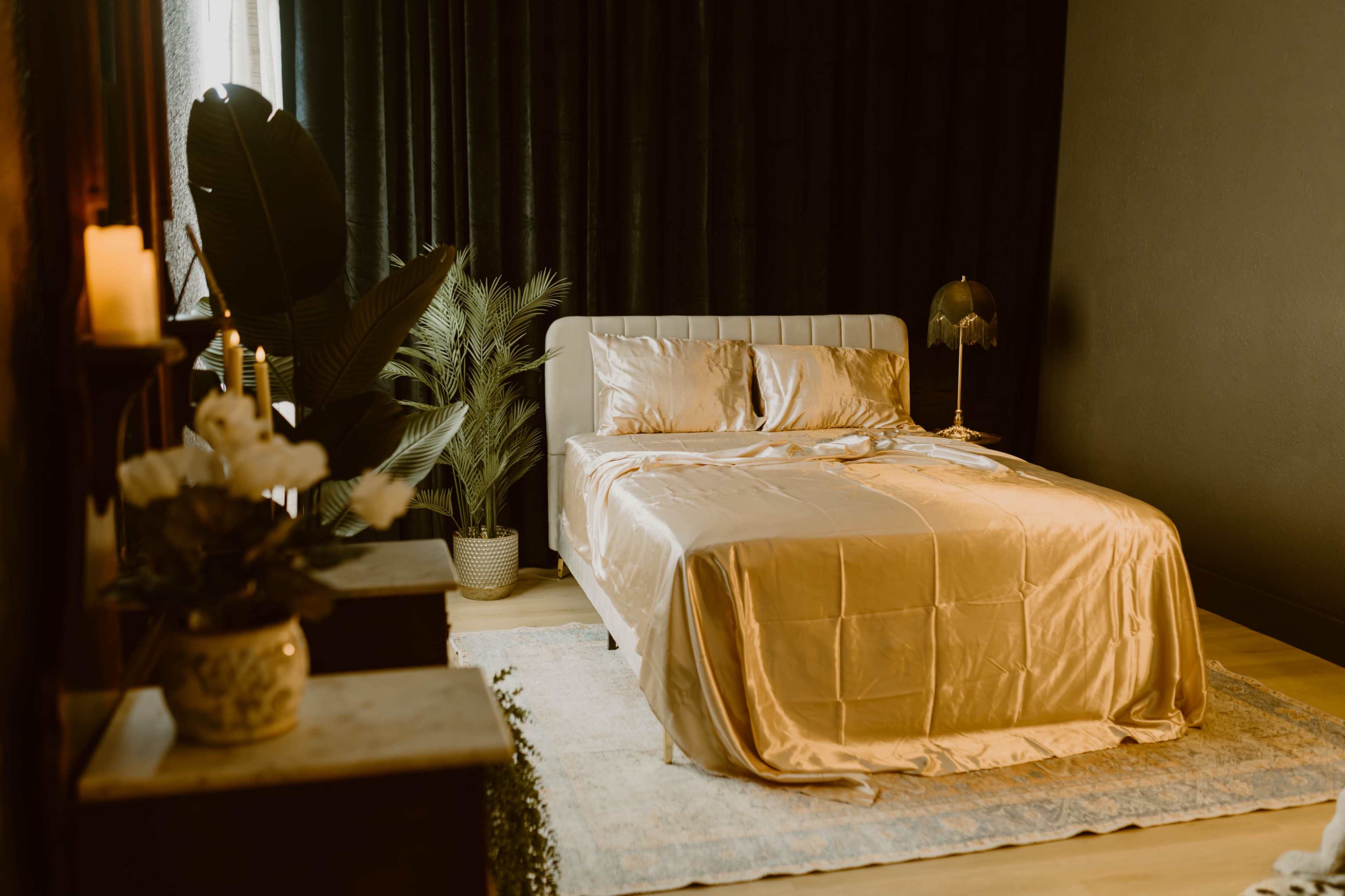 A neatly made bed with gold bedding is positioned in a dark room adorned with green plants and soft lighting.
