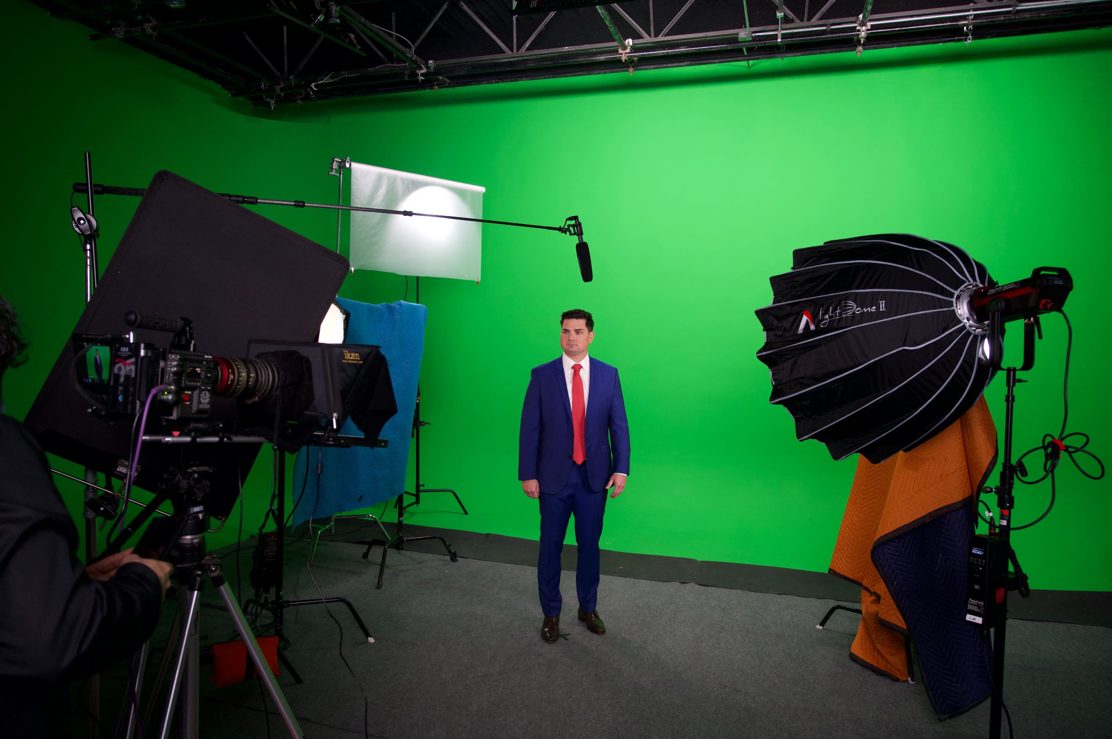 A man in a blue suit and red tie stands in front of a green screen in a photography studio, surrounded by lighting and equipment.
