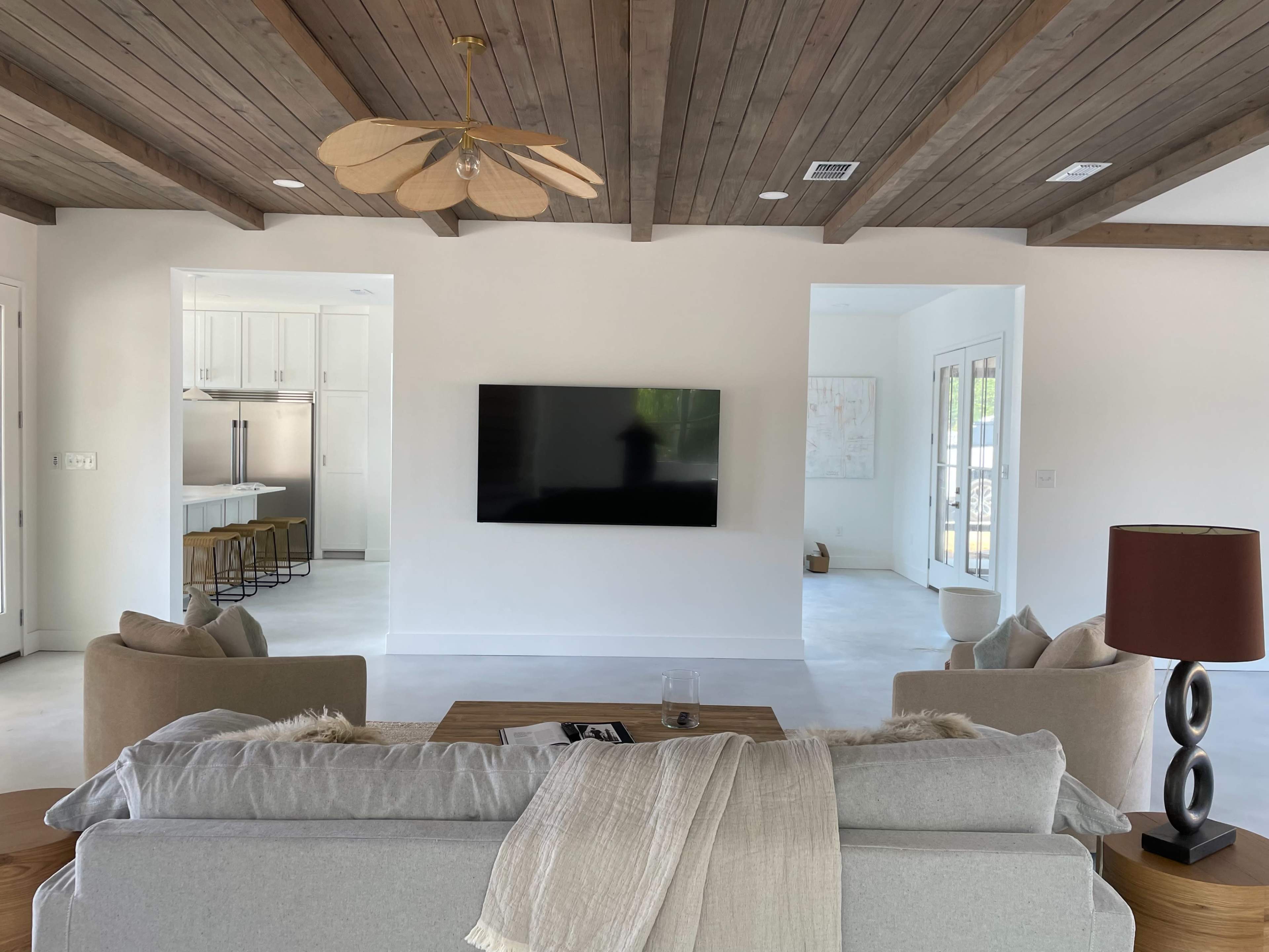 A modern living room with a sofa facing a mounted flat-screen television, flanked by two armchairs, and featuring light-colored walls and wooden ceiling beams.