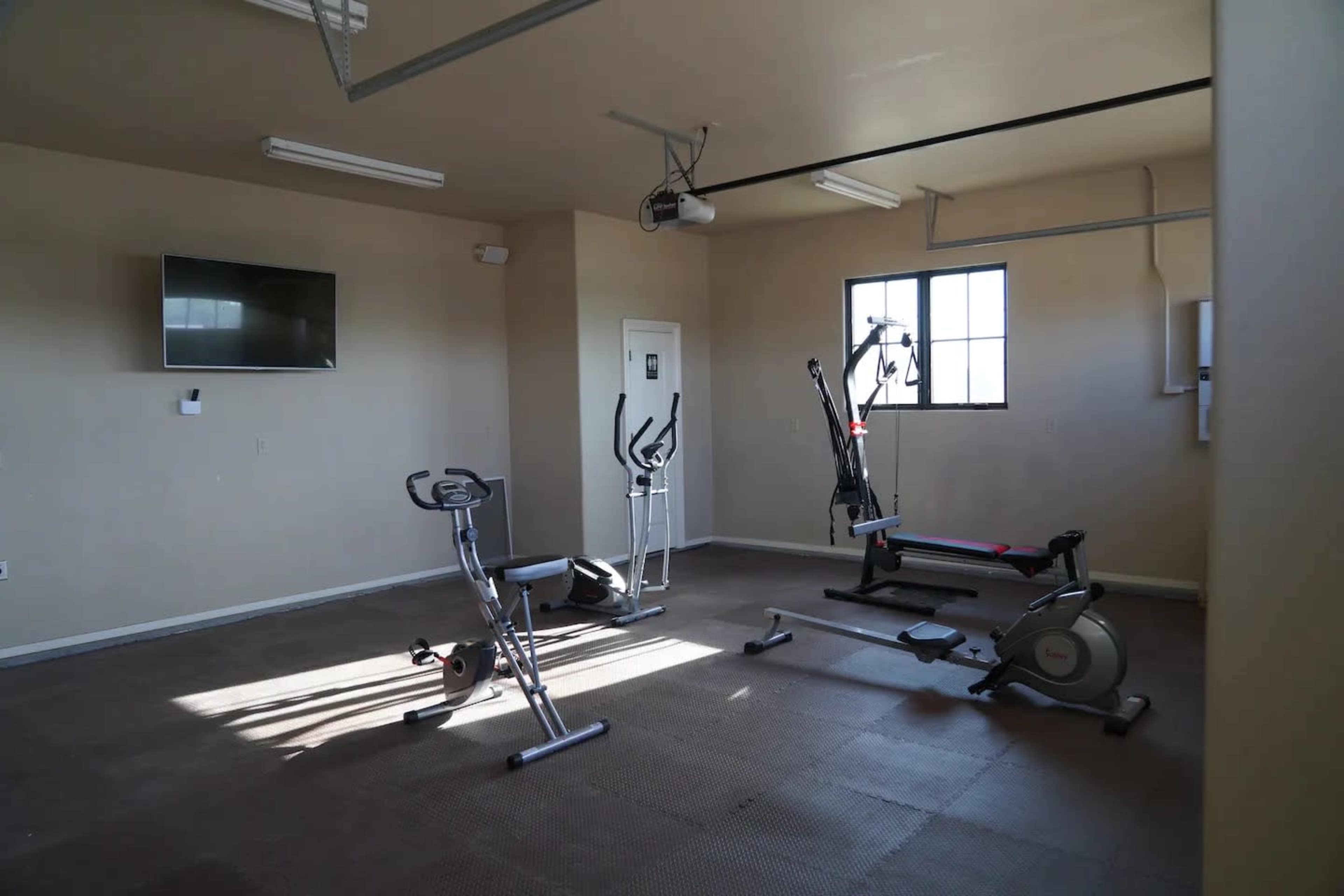 A gym room with exercise equipment including a stationary bike, elliptical machine, and weight bench, along with a wall-mounted television and a window.