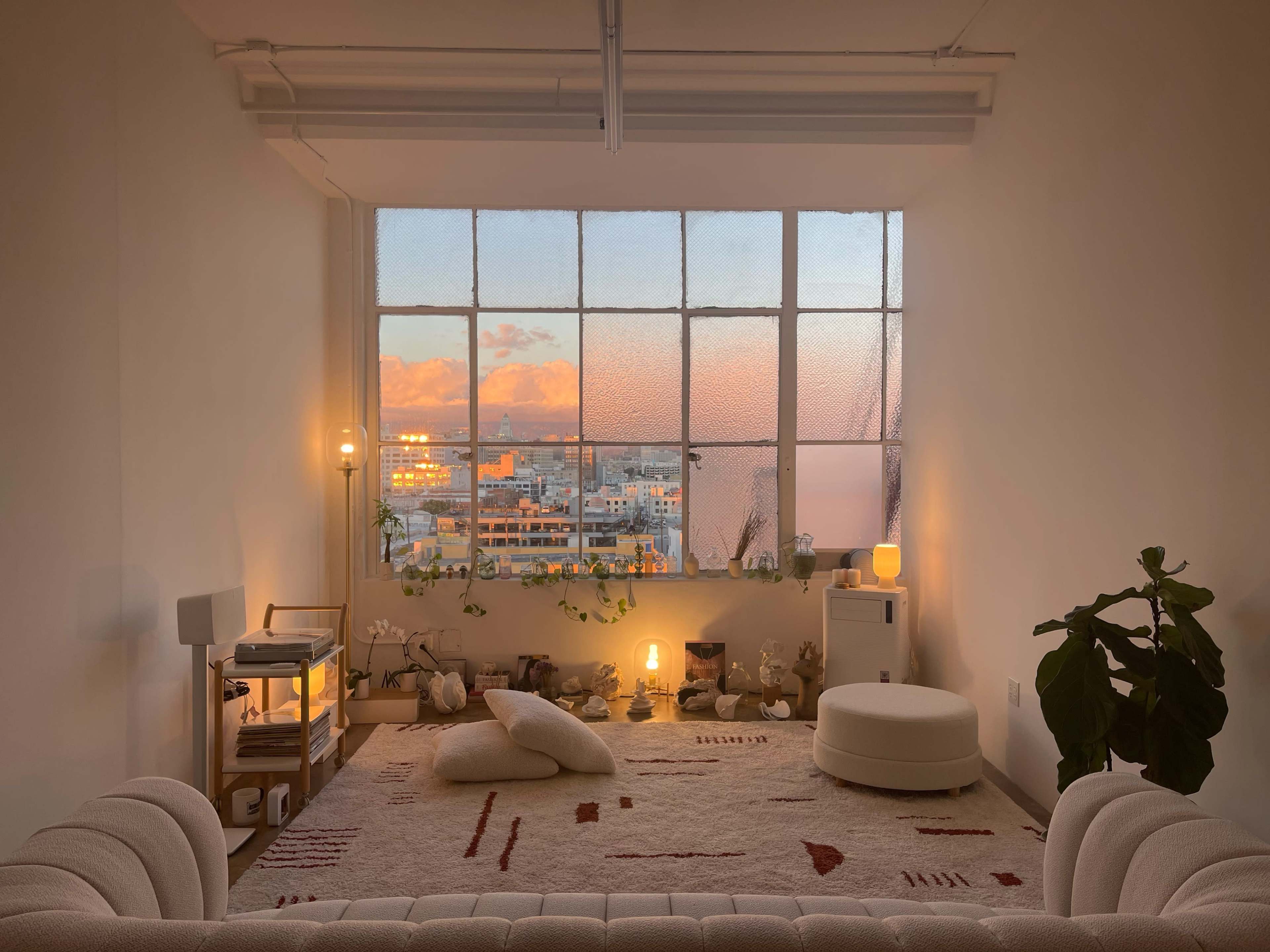 A cozy interior with a large window revealing a sunset view, featuring minimalistic furniture and plants.