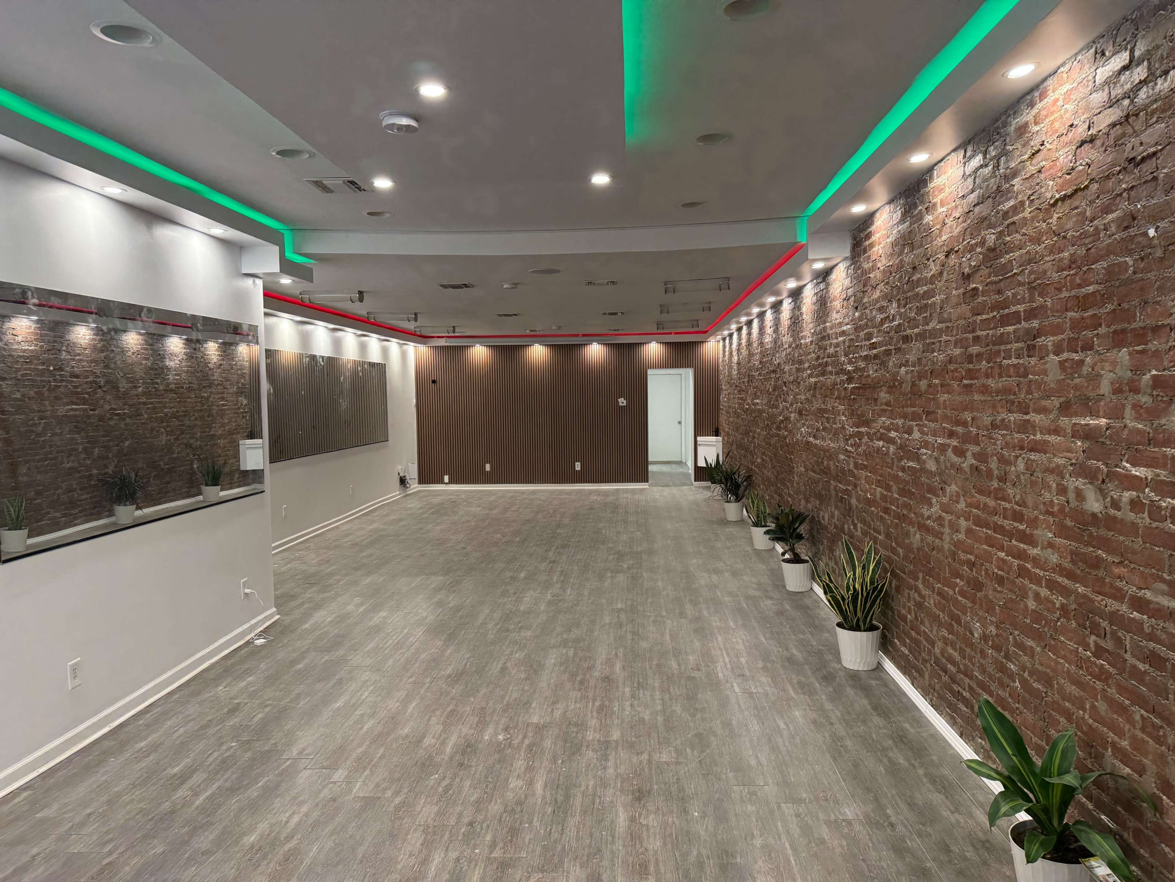 The image shows a spacious interior room with exposed brick walls, a mirrored section, and green and red LED lighting along the ceiling, complemented by potted plants along the floor.