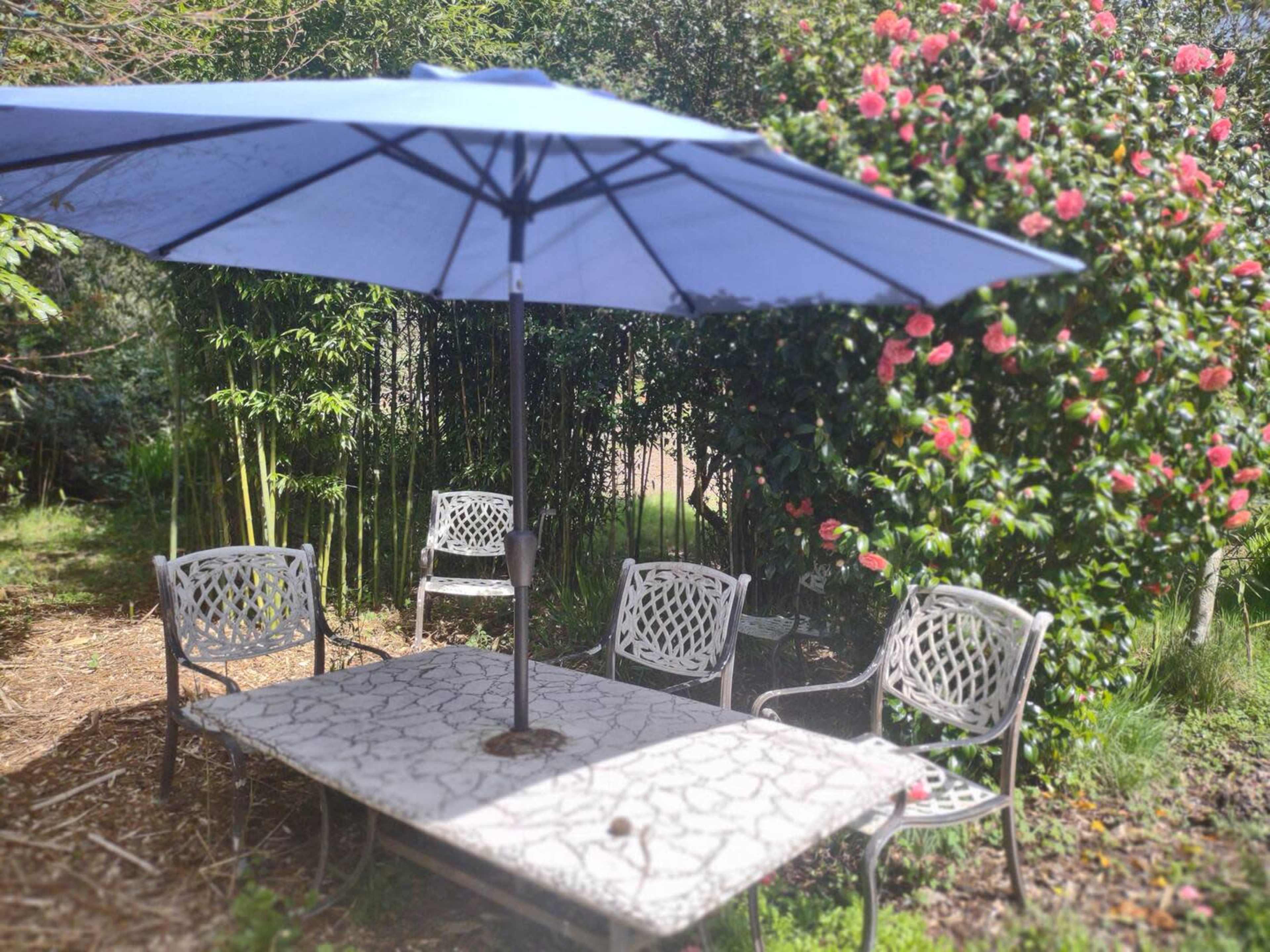 A blue umbrella shades a metal table surrounded by four chairs in a garden with blooming bushes.