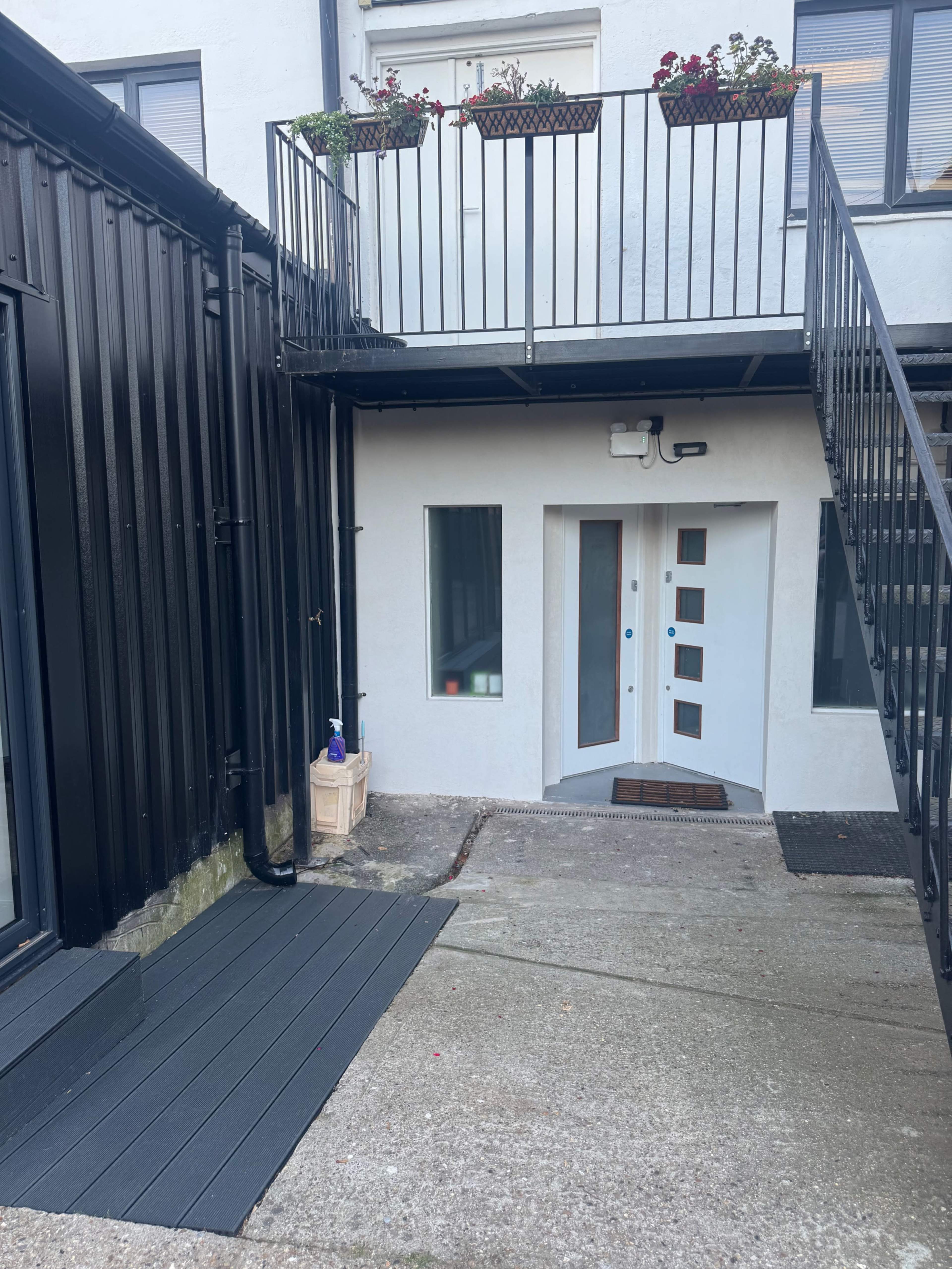 The image shows a small outdoor area with a stairway leading to a balcony and an entrance featuring a modern white door with geometric windows.