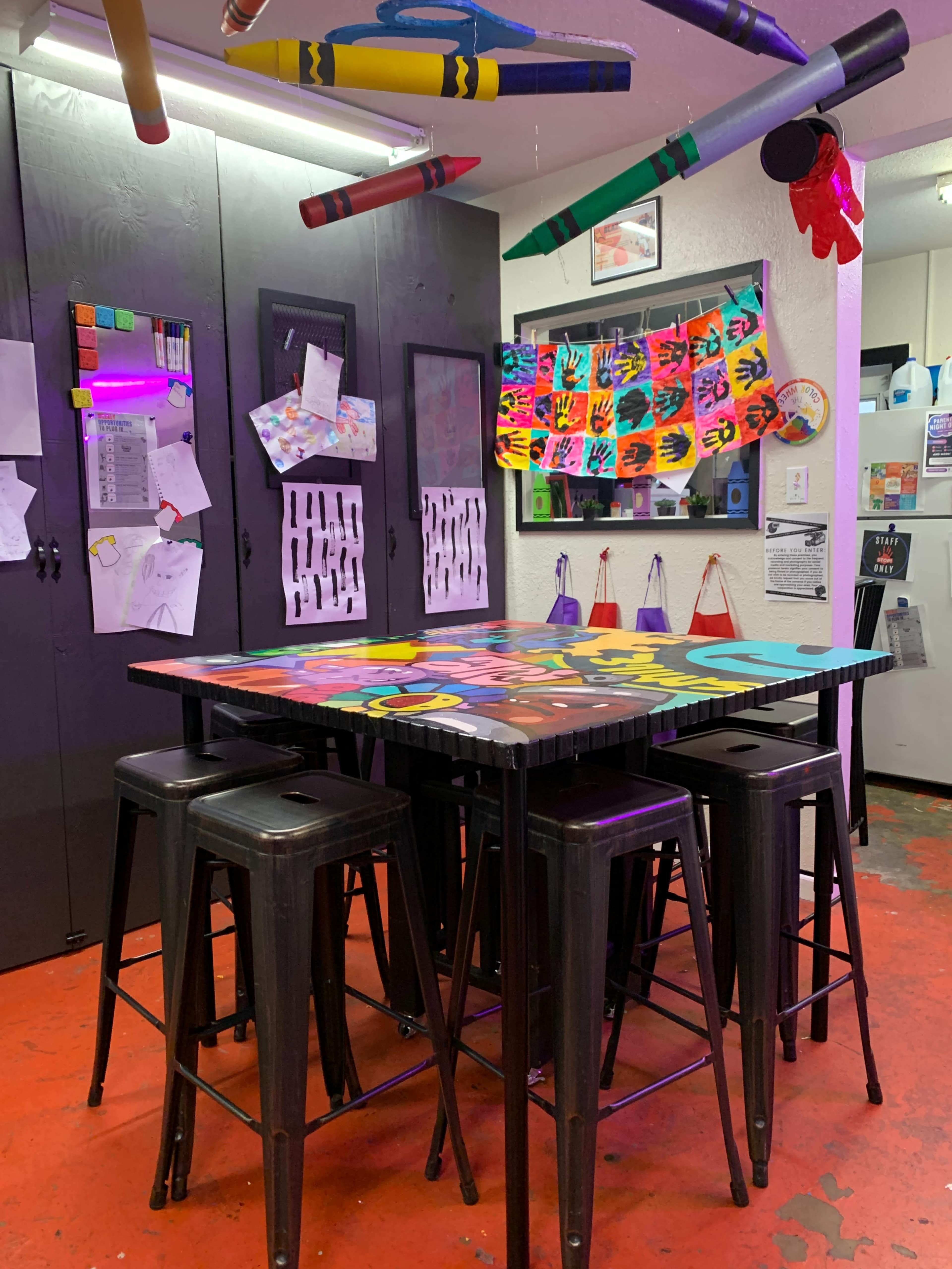 A colorful table surrounded by metal stools is situated in a room decorated with drawings, crafts, and art supplies hanging on the walls.
