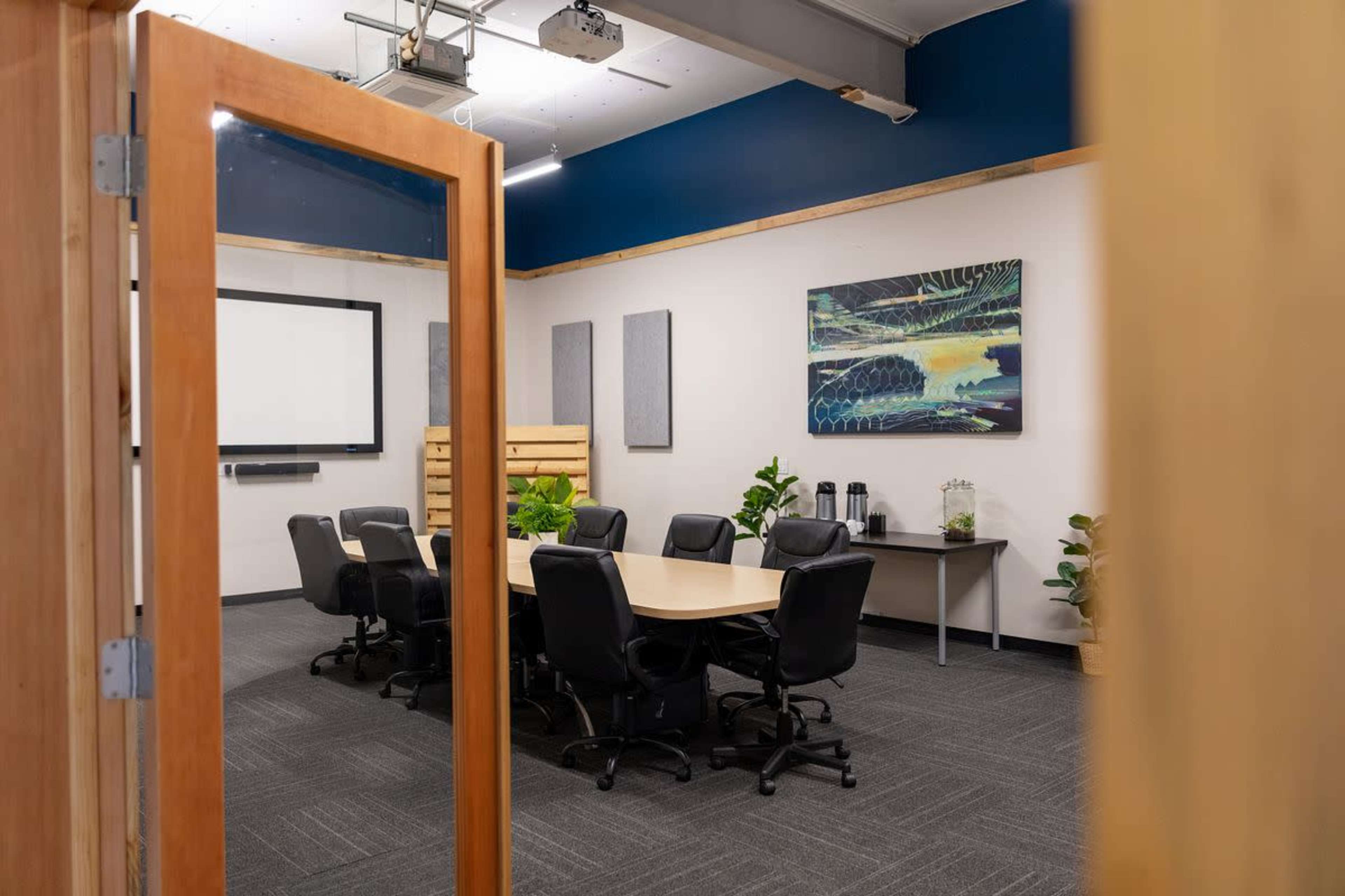 A conference room with a round table surrounded by black chairs, featuring a large screen and plant decorations, is visible through a doorway.