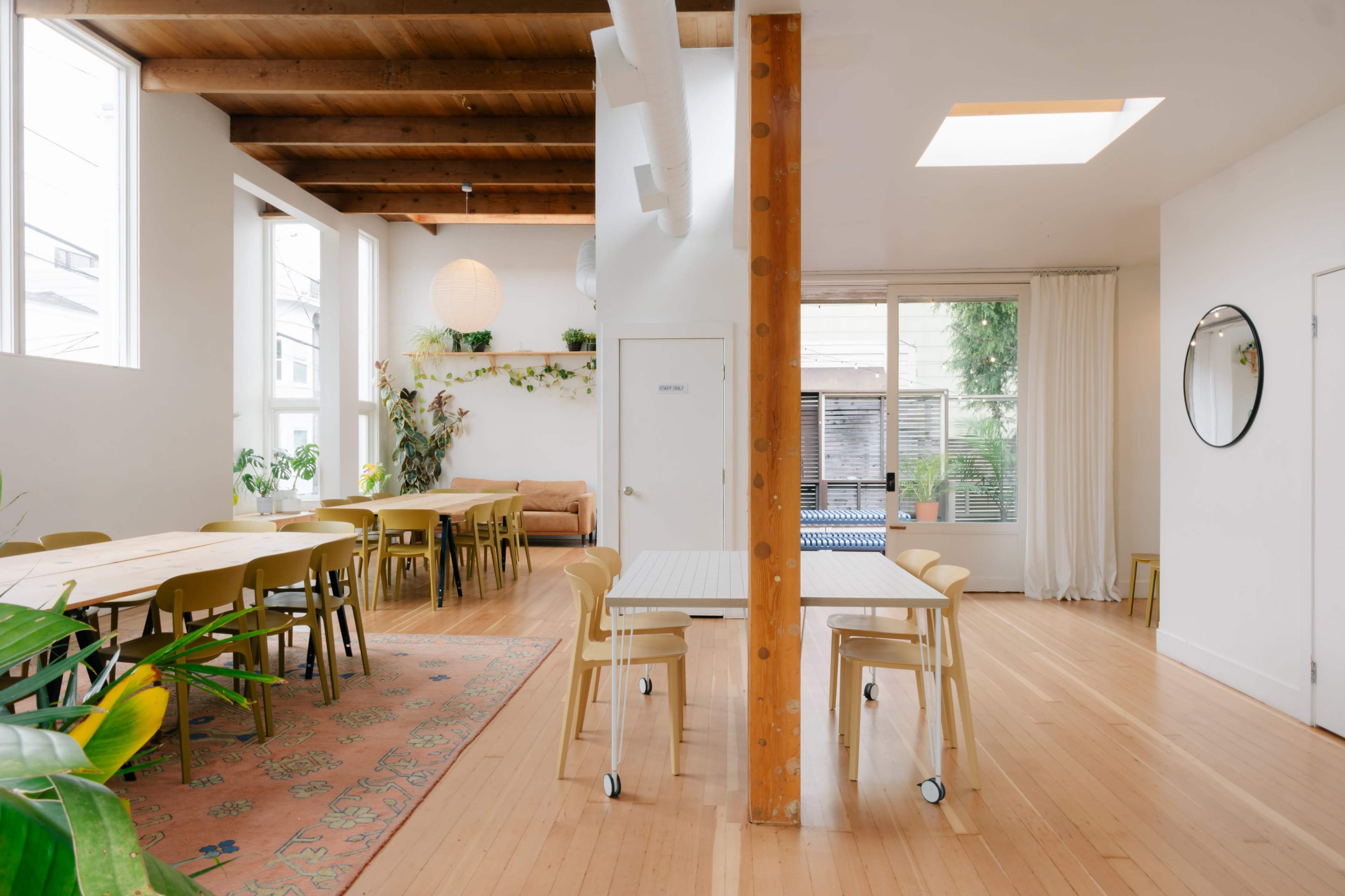 The image shows a bright, airy interior space with wooden beams, a mix of seating areas, and large windows, creating a welcoming atmosphere.