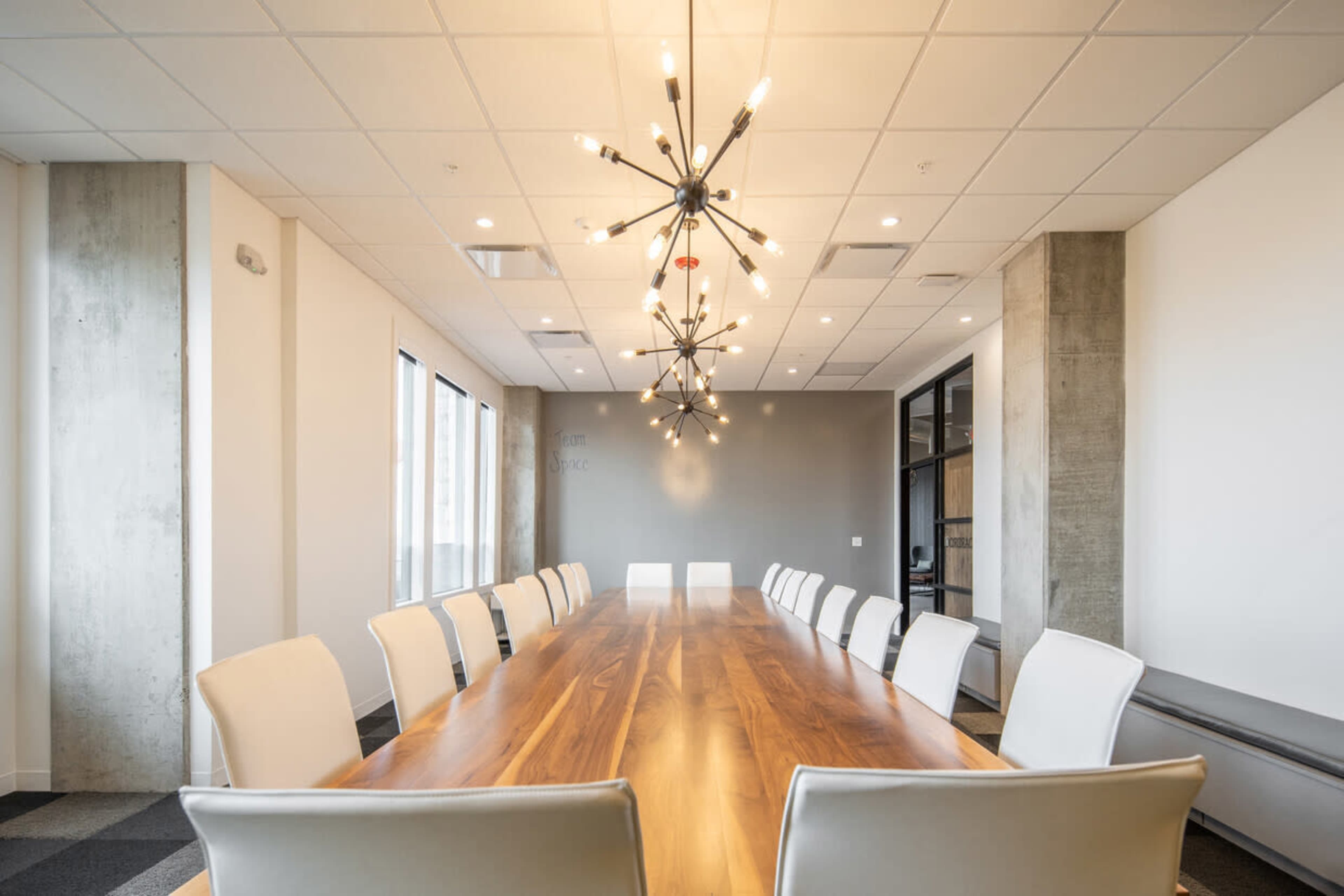 A long wooden conference table with white chairs is situated in a well-lit meeting room featuring modern chandeliers.