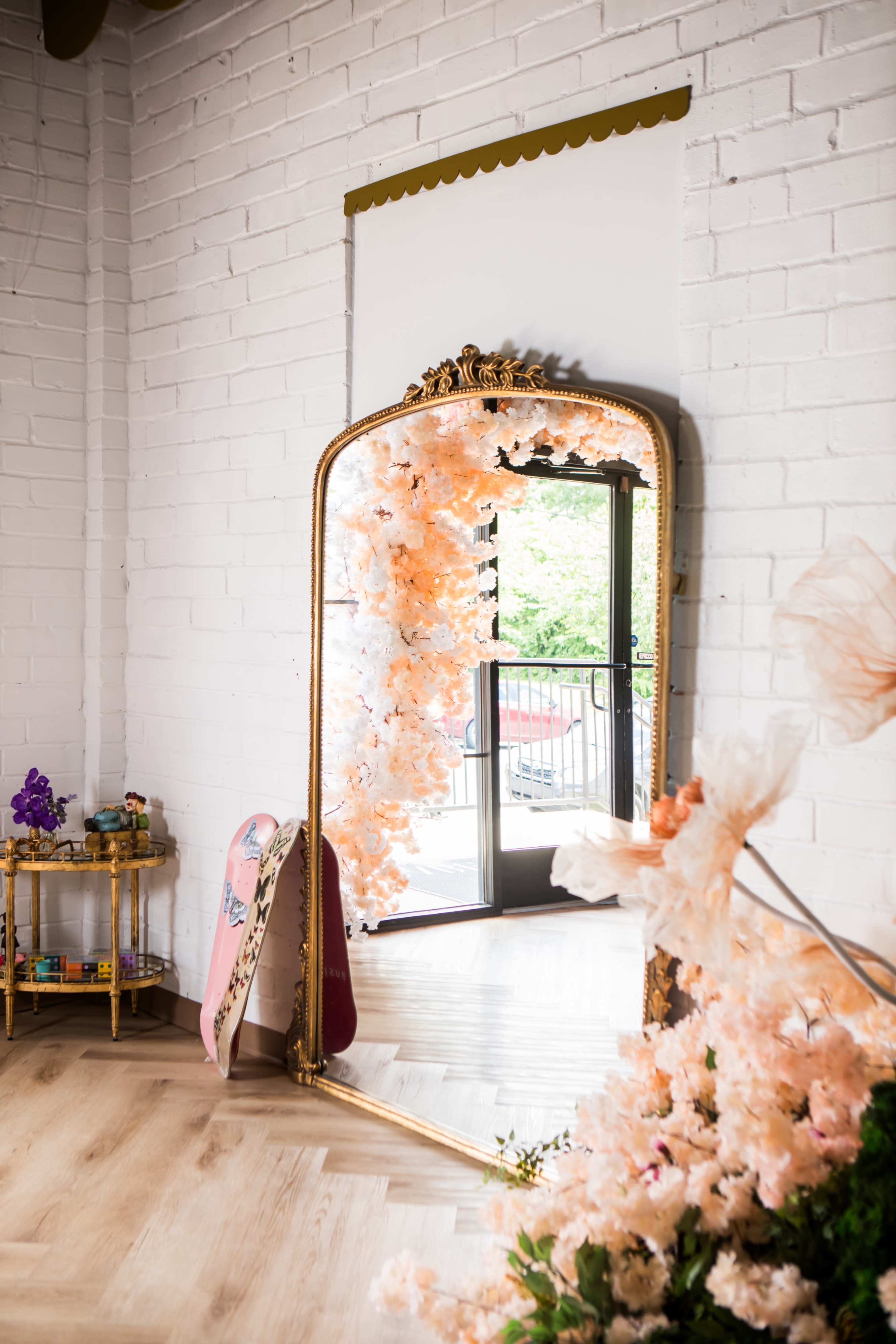 A large ornate mirror with a floral arrangement in shades of peach frames an open door leading outside in a bright interior space.