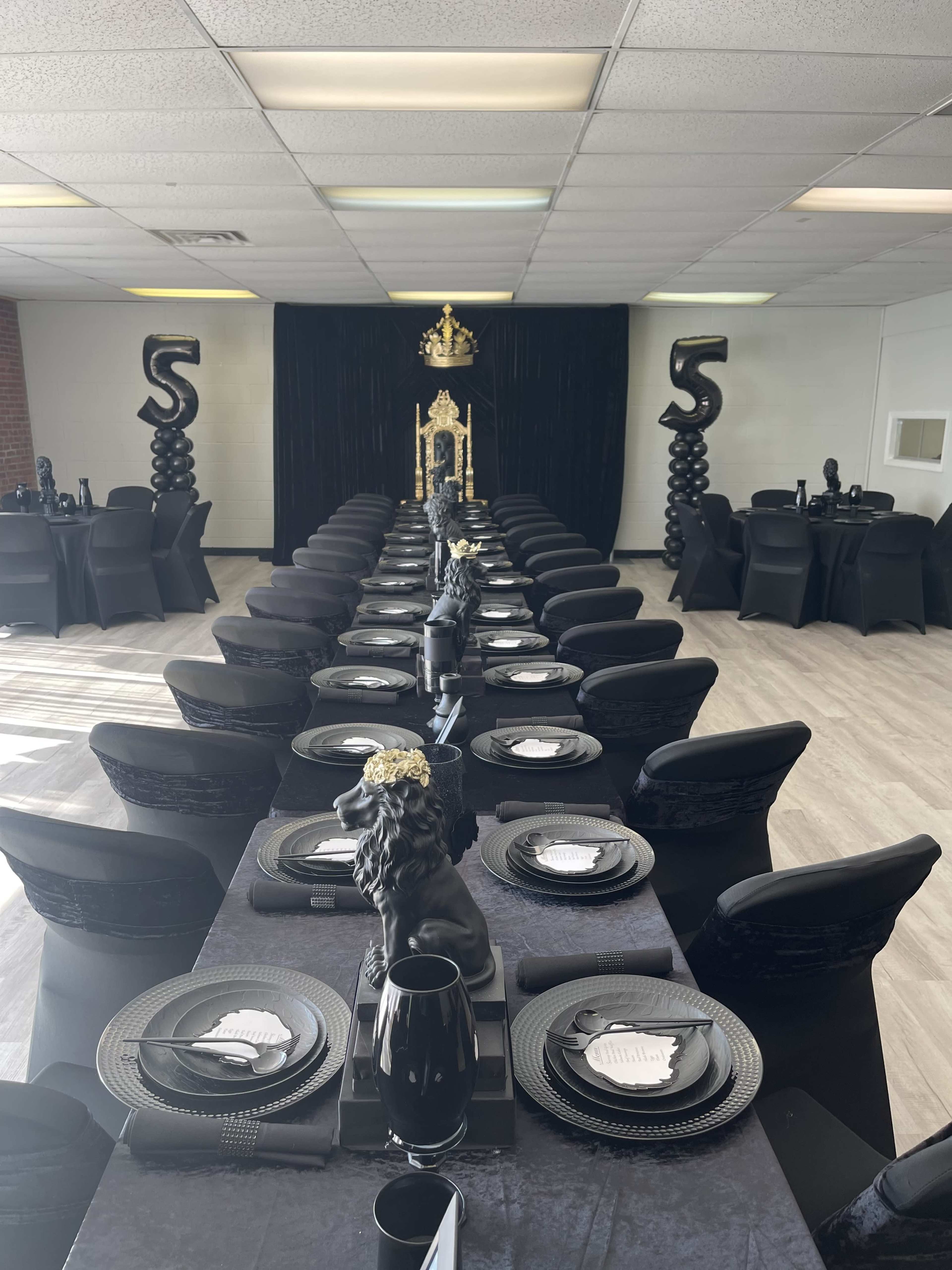 A long rectangular dining table is set with black tableware and decorated with lion statues, surrounded by chairs, in a spacious, elegantly arranged event room.